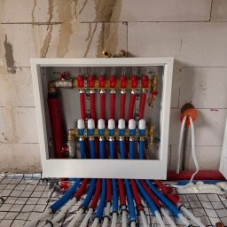 BartSystem Bartosz Kaczmarski - Rozdzielacz ogrzewania podłogowego z rurami w kolorach czerwonym i niebieskim, zamontowany na ścianie z bloczków, z widoczną folią izolacyjną na podłodze.