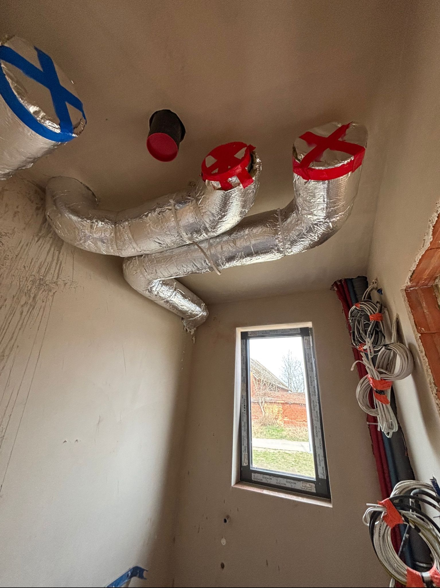 Instalacja wentylacyjna z rurami aluminiowymi pod sufitem, zabezpieczonymi taśmą. W tle okno i zwisające kable, ściany w trakcie wykończenia.