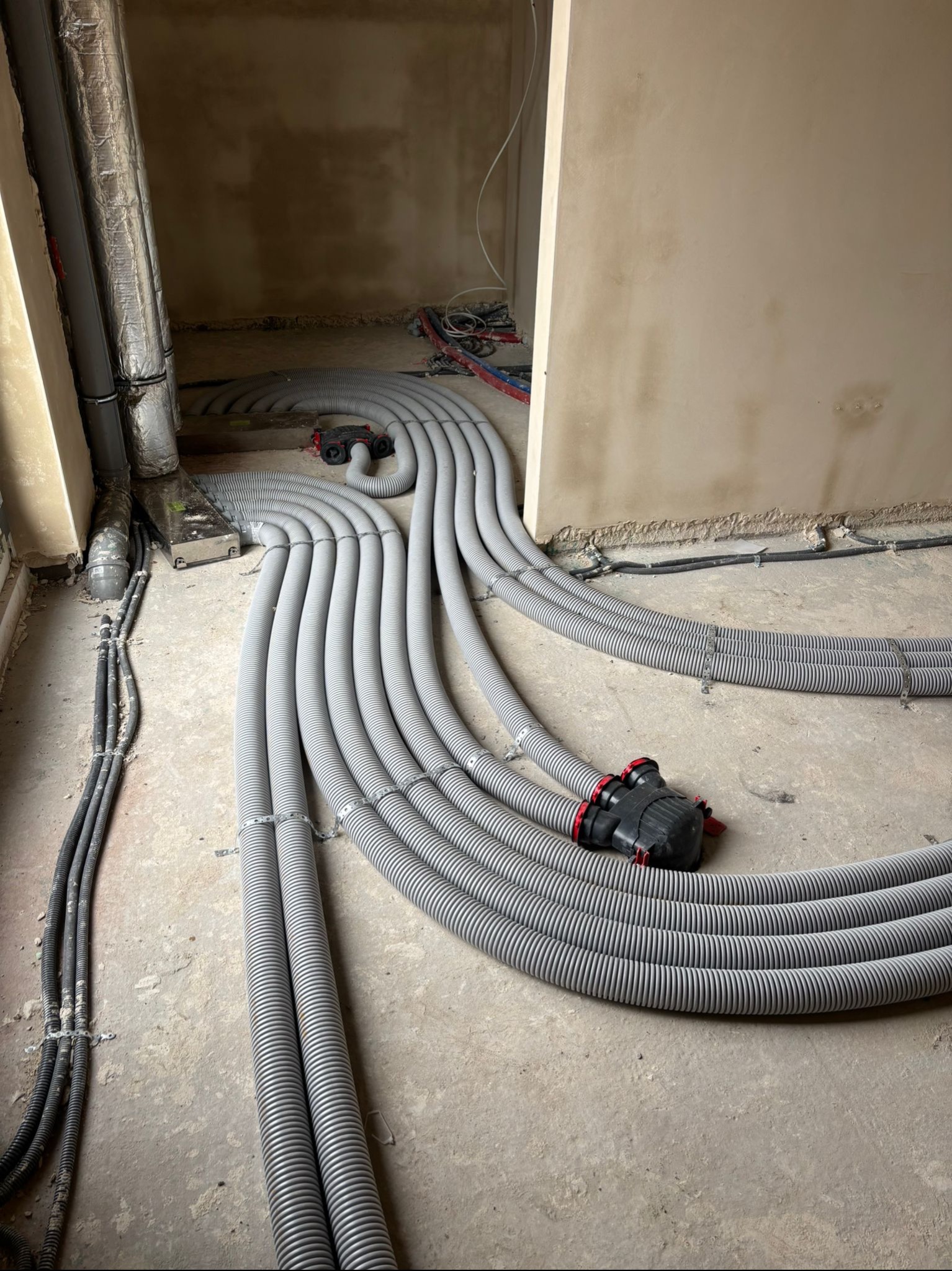 Szare, elastyczne rury wentylacyjne ułożone na surowym podłożu wewnątrz budynku, z widocznymi elementami instalacji elektrycznej i wentylacyjnej w tle.