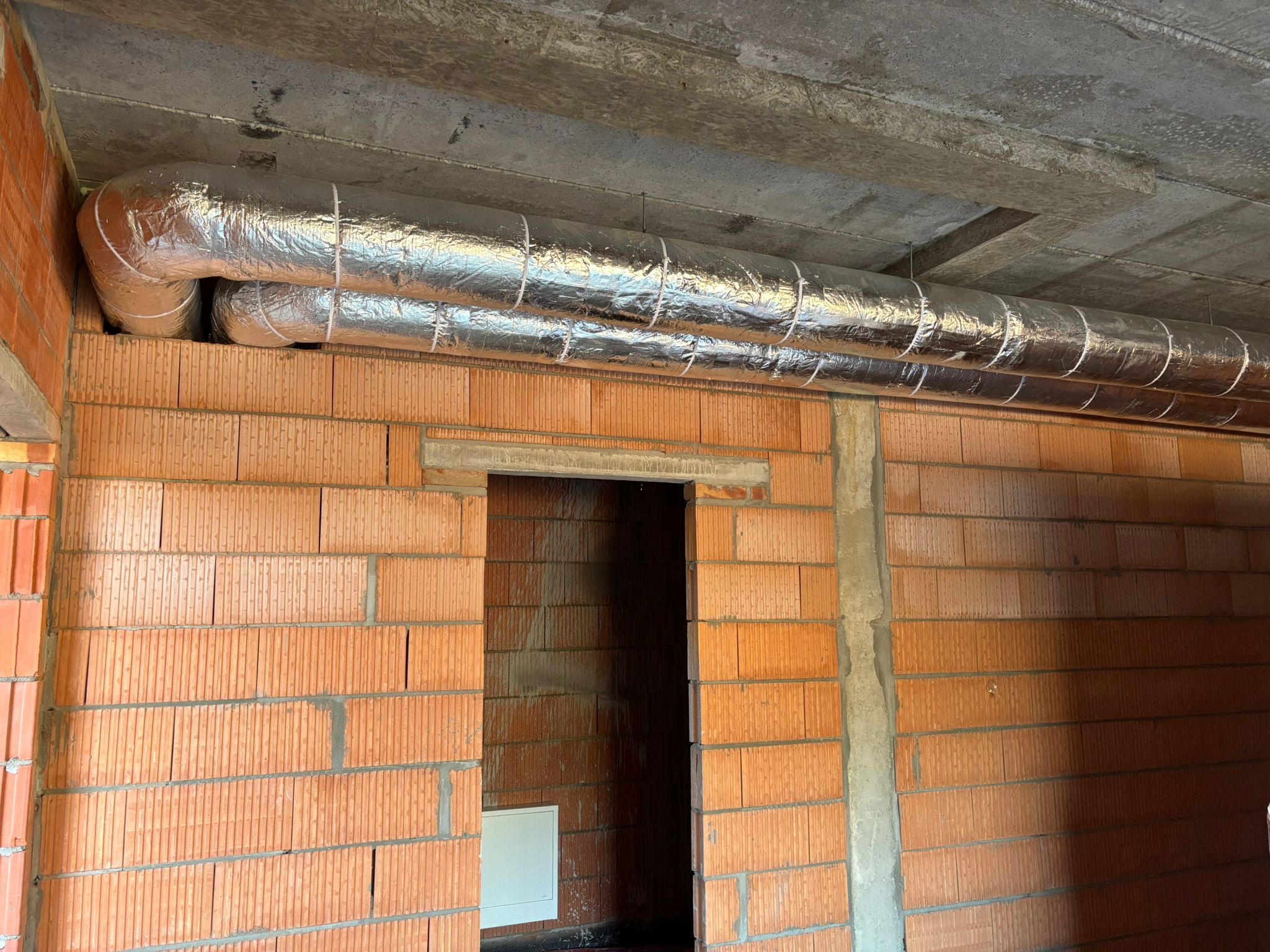 Dwa izolowane przewody wentylacyjne z folii aluminiowej przymocowane do sufitu w surowym wnętrzu z ceglanymi ścianami i otwartym przejściem.