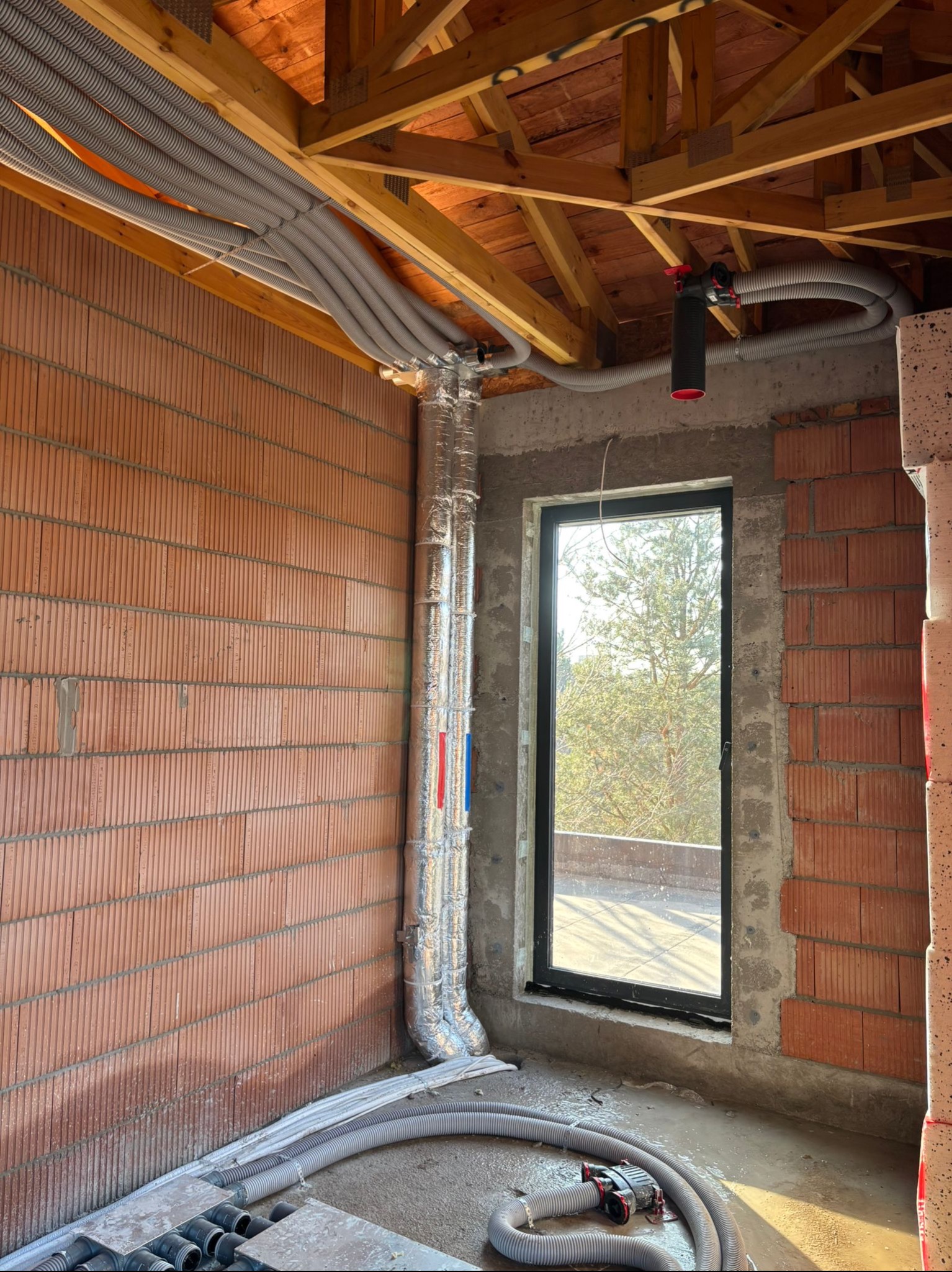Wnętrze w trakcie instalacji rekuperacji: widoczne przewody wentylacyjne pod stropem, elementy instalacji na podłodze, surowa cegła i okno z widokiem na zewnątrz.