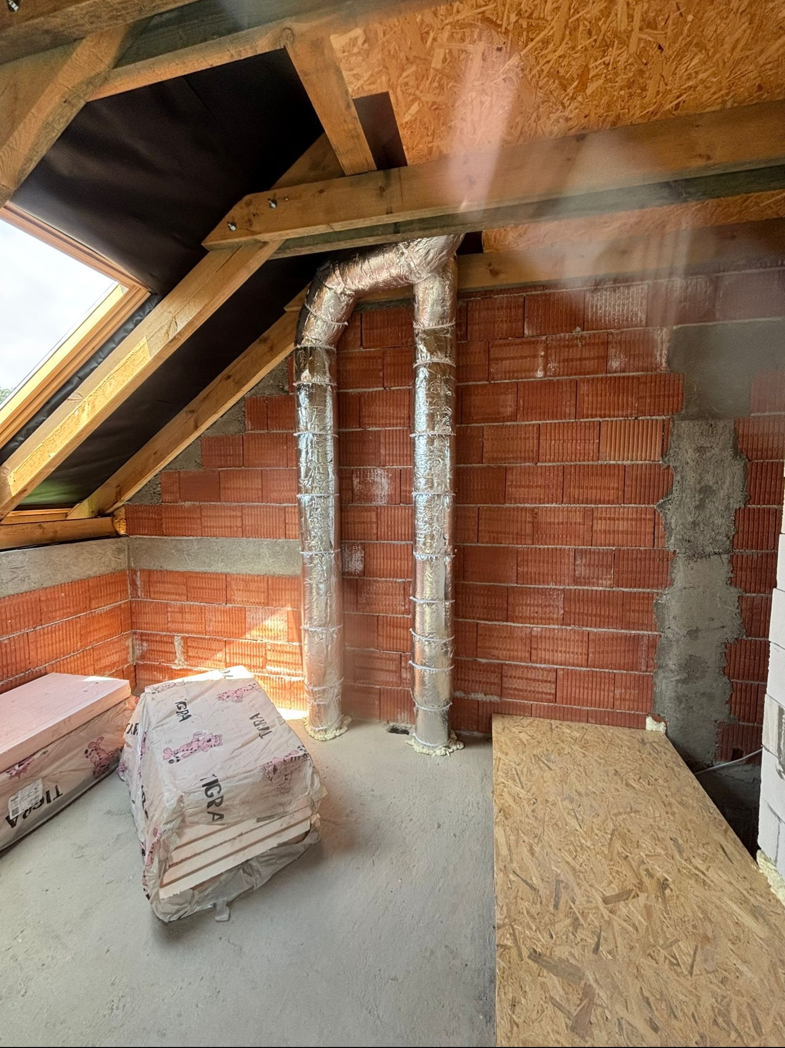 Montaż instalacji wentylacyjnej: Dwie srebrne rury wentylacyjne w folii aluminiowej wznoszą się pionowo w surowym wnętrzu poddasza z oknem dachowym i czerwoną cegłą.