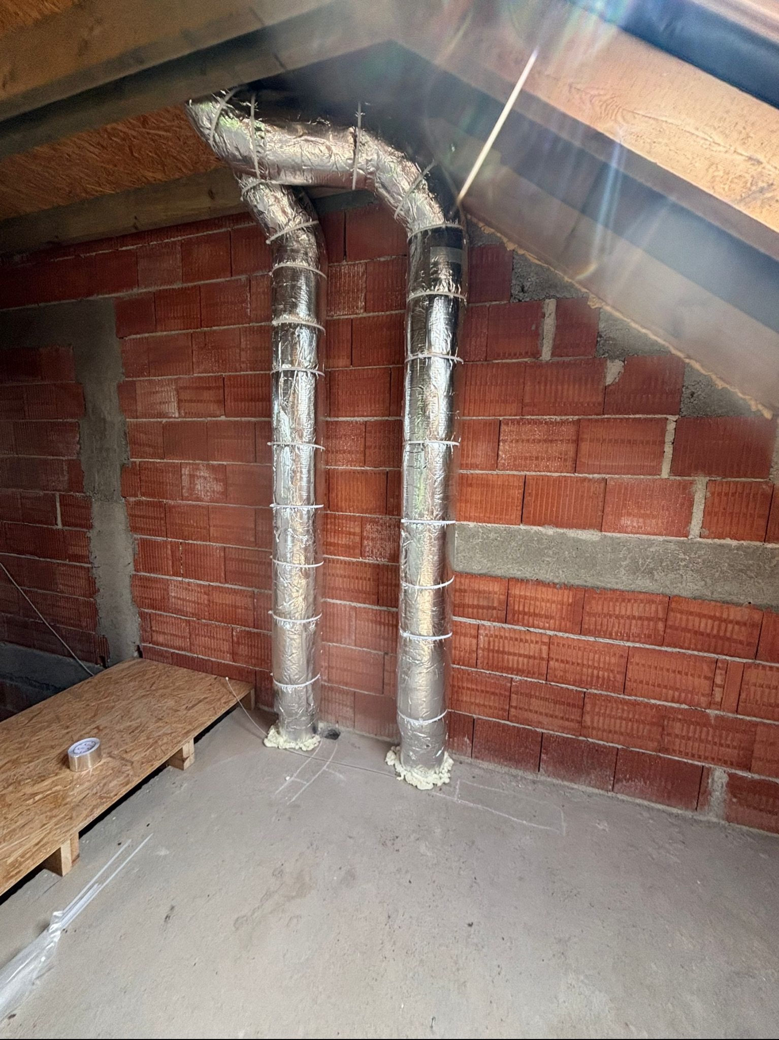 Dwa pionowe przewody wentylacyjne w izolacji, zamocowane do ściany z czerwonej cegły w surowym wnętrzu poddasza. Widoczne elementy konstrukcji dachu i podłogi.