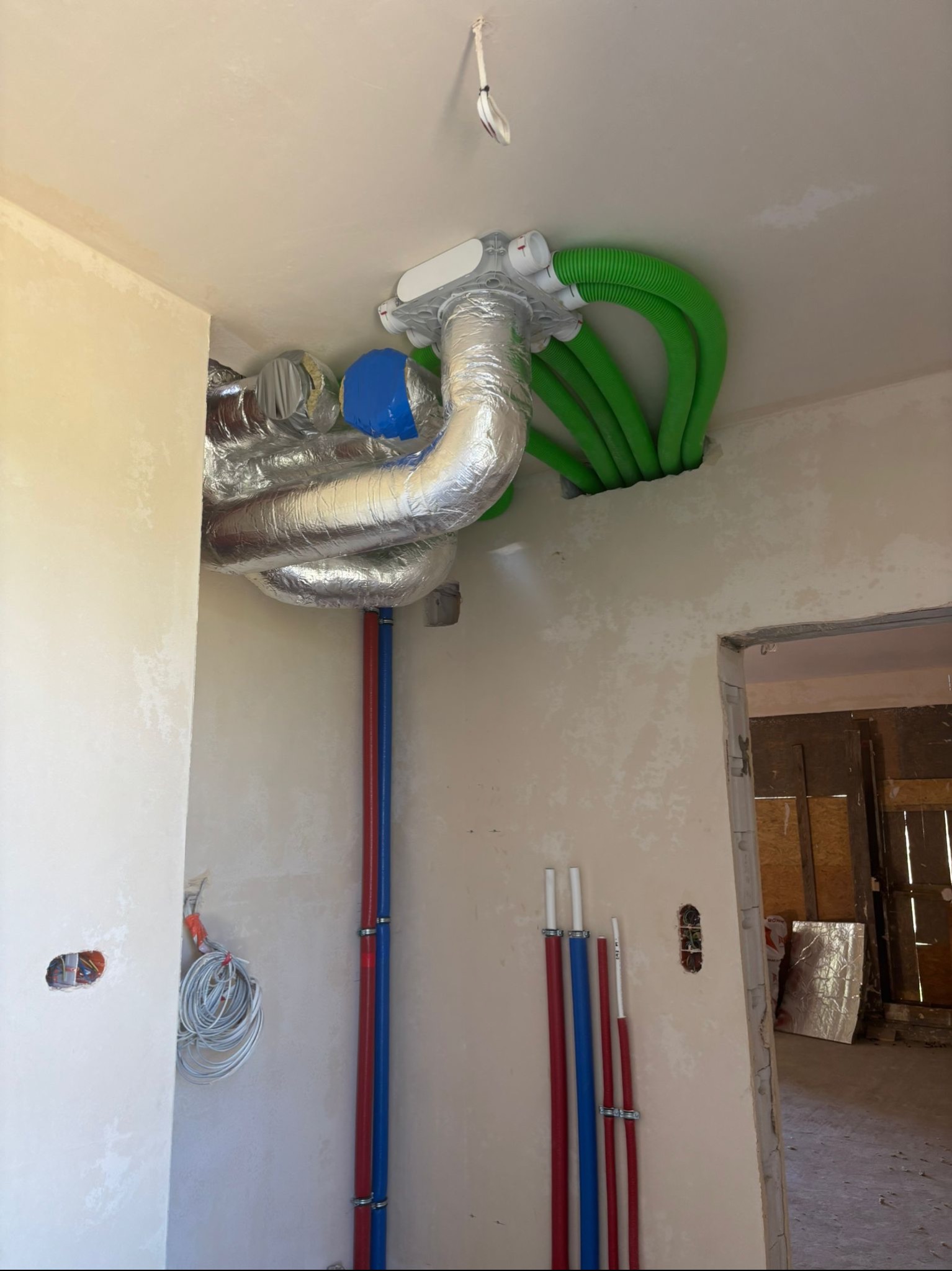 Instalacja rekuperacji z zielonymi rurami wentylacyjnymi, srebrne rury izolowane, oraz pionowe rury wodne w kolorze niebieskim i czerwonym, w surowym wnętrzu.