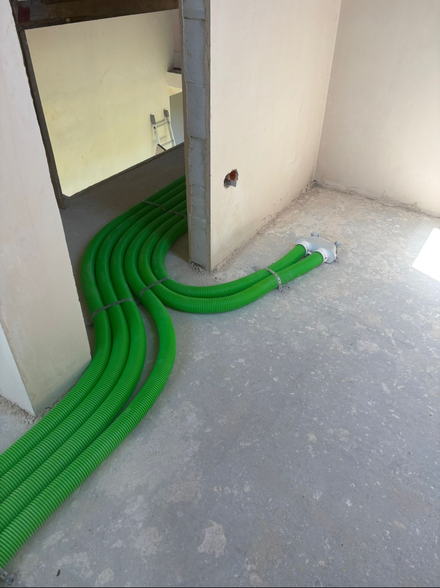 Zielone rury wentylacyjne prowadzone wzdłuż ściany w surowym wnętrzu. Widoczna drabina w tle. Instalacja przed wylaniem posadzki.