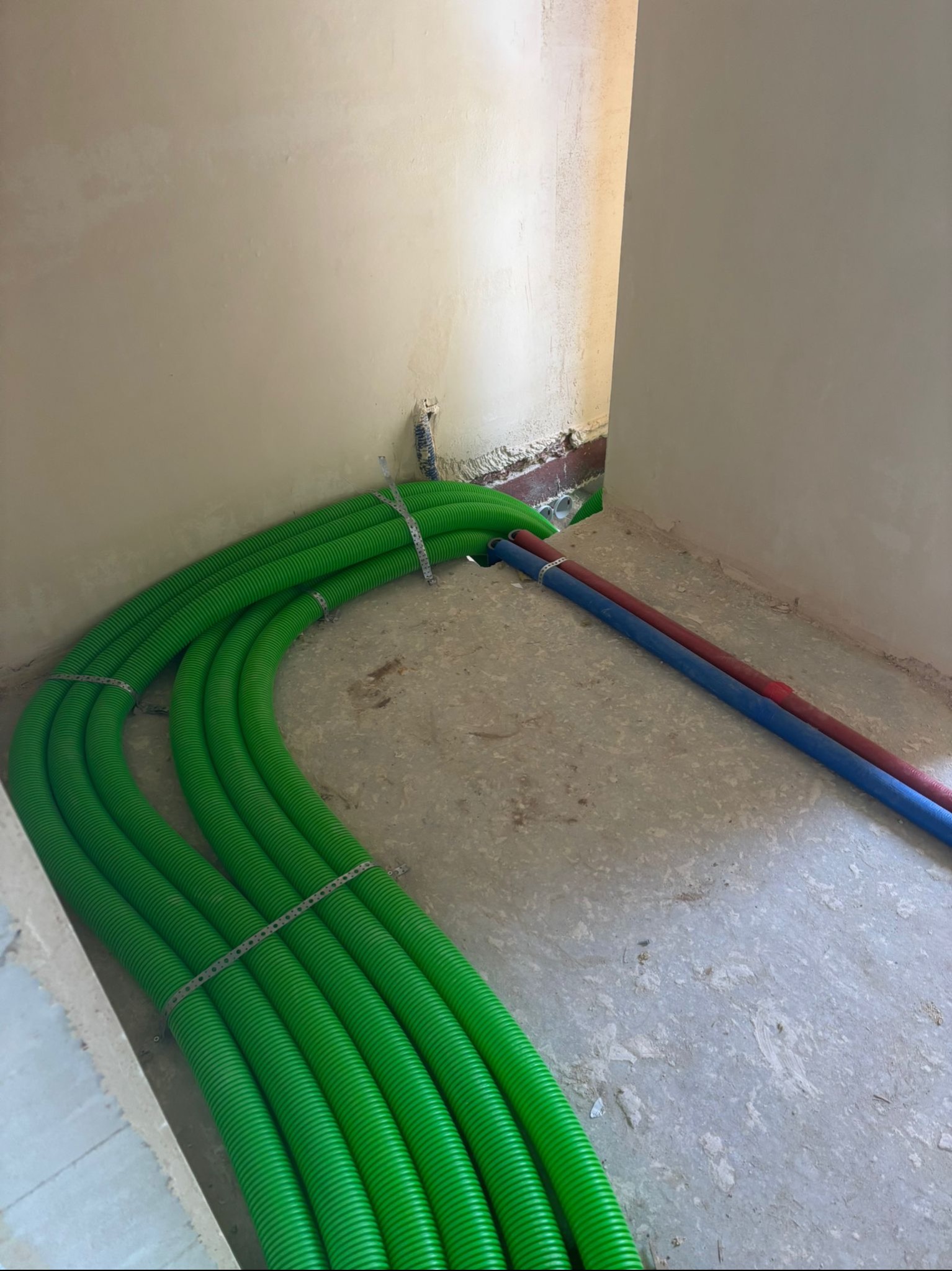 Zielone rury podłogowe, spięte metalową taśmą, układane na surowym betonie w rogu pomieszczenia, z widocznymi fragmentami rur czerwonych i niebieskich.