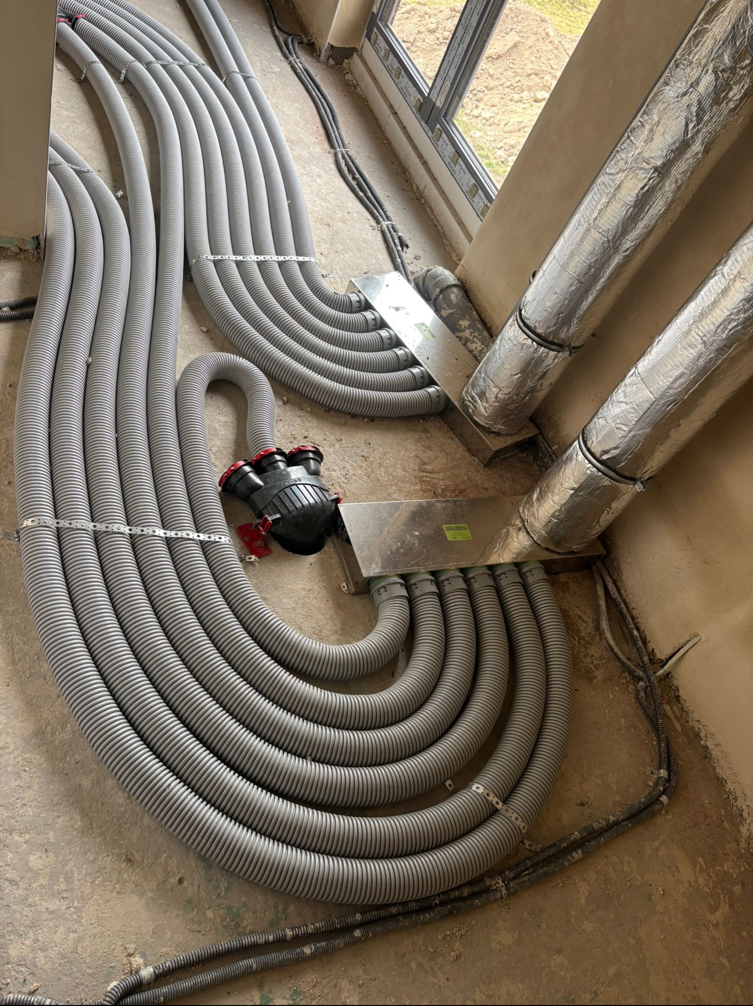 Szare, karbowane rury wentylacyjne ułożone na surowej podłodze w budynku w stanie deweloperskim, prowadzące do srebrnych, zaizolowanych przewodów wentylacyjnych.