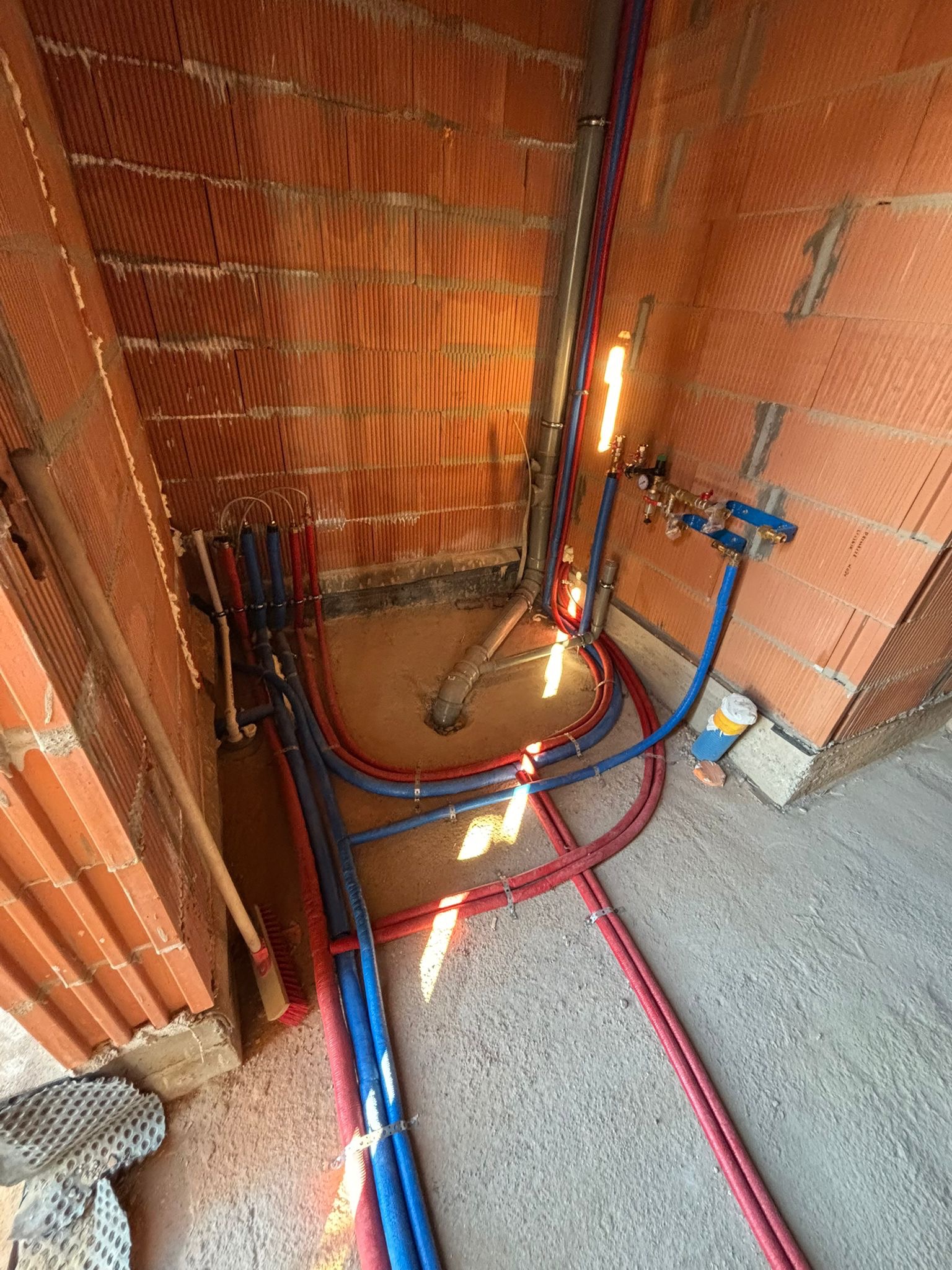 Instalacja rur grzewczych (czerwone i niebieskie) w surowym wnętrzu z ceglanymi ścianami. Widoczne przyłącza i fragment podłogi. Prace hydrauliczne w trakcie realizacji.
