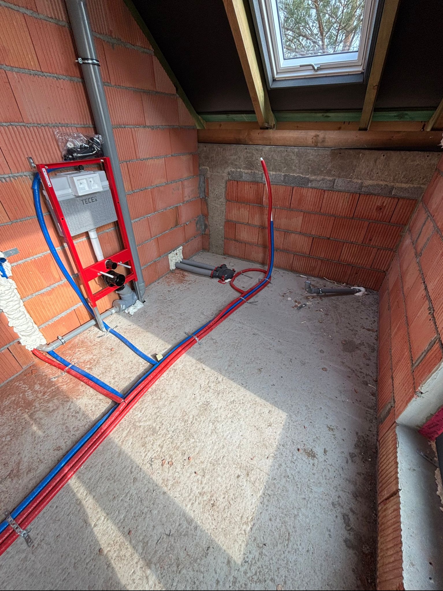 Surowy stan łazienki na poddaszu z widoczną instalacją wodno-kanalizacyjną: stelaż WC Tece, rury PEX w kolorze czerwonym i niebieskim, okno dachowe.