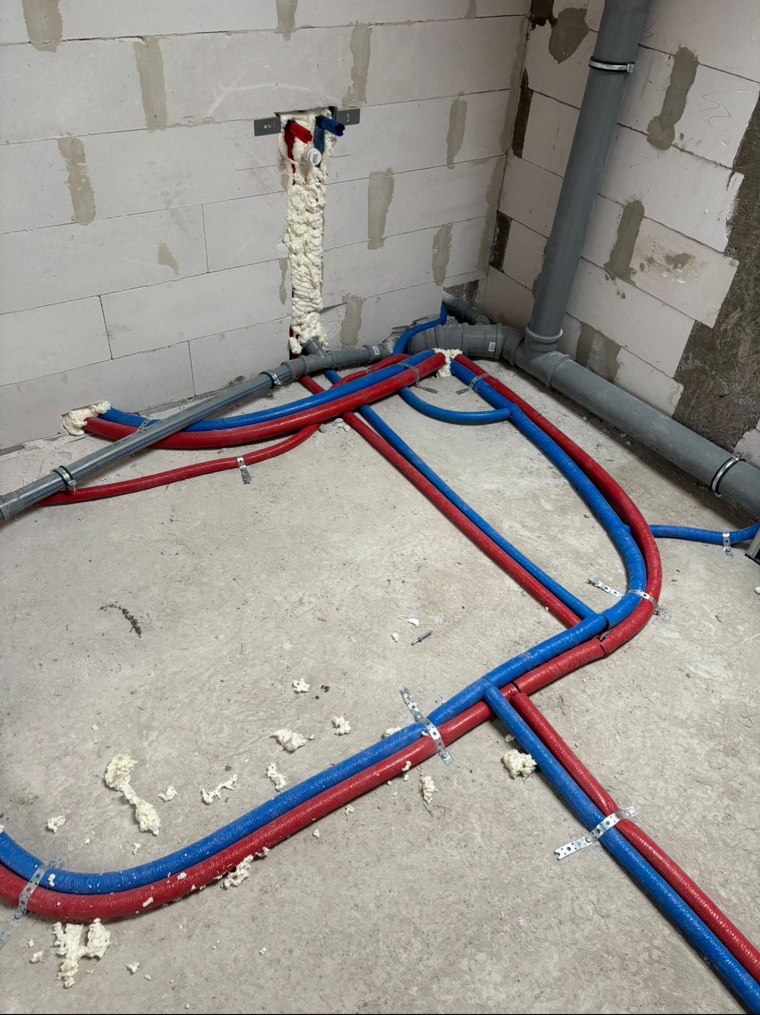 Instalacja rur grzewczych (czerwone i niebieskie) w surowym stanie, z pianką montażową przy ścianach z bloczków. Szare rury kanalizacyjne w tle.