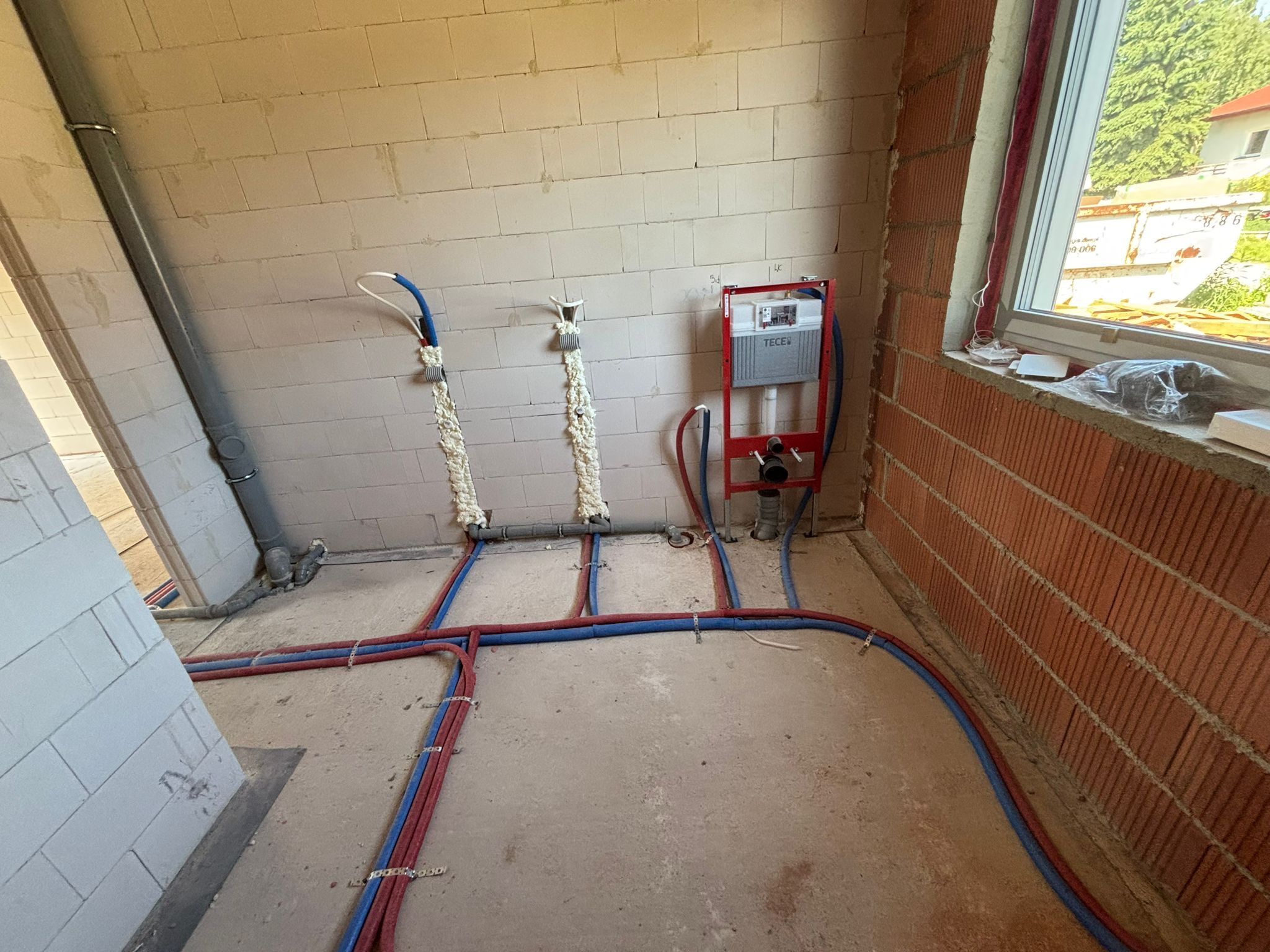 Instalacja wodno-kanalizacyjna w surowym wnętrzu: rury w kolorach czerwonym i niebieskim prowadzone po podłodze, zamocowane do ściany z pustaków, widoczna spłuczka TECE.