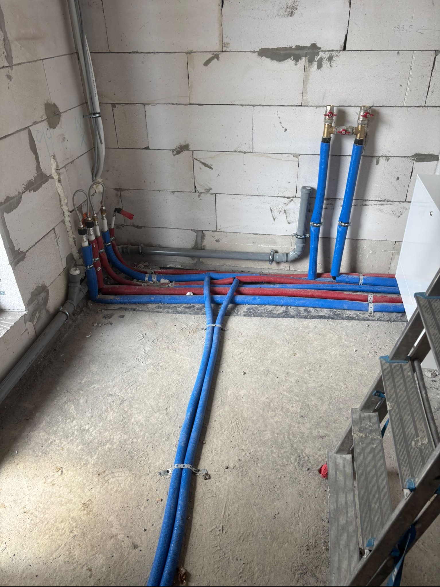 Instalacja wodna w surowym wnętrzu: niebieskie i czerwone rury prowadzone wzdłuż ściany z bloczków, widoczne zawory i drabina. Etap prac instalacyjnych.