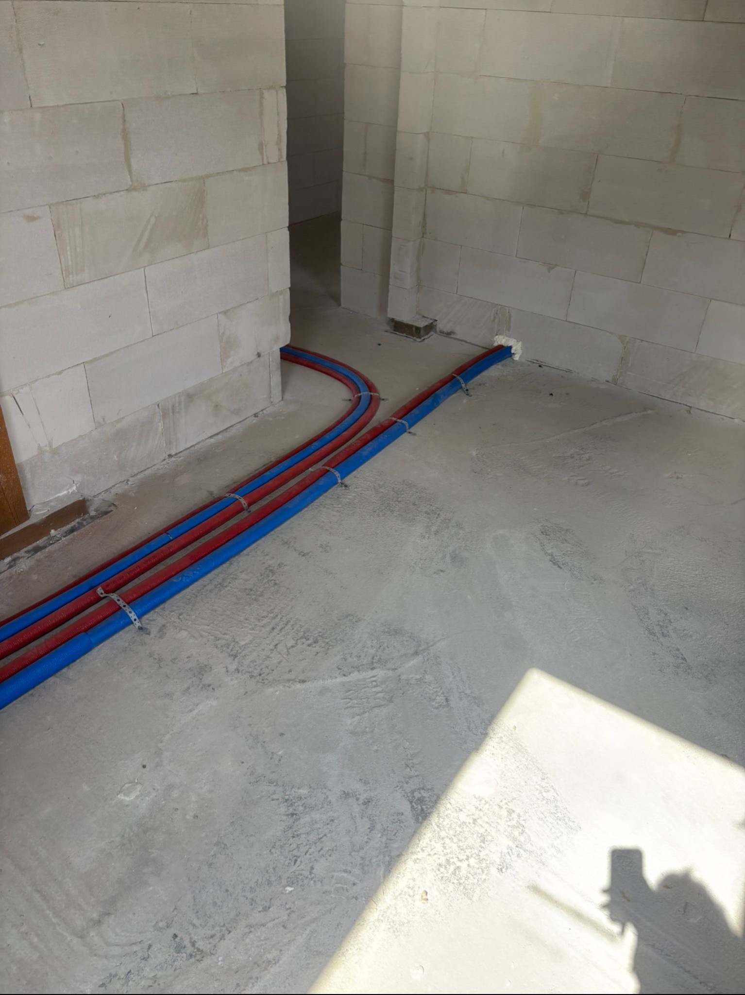 Instalacja ogrzewania podłogowego: czerwone i niebieskie rury, przymocowane do podłoża, prowadzone wzdłuż ściany z białego betonu komórkowego. Widoczny cień osoby.