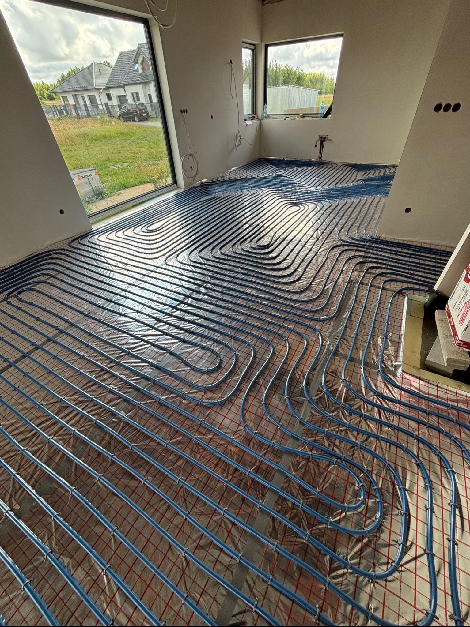 Niebieskie rury ogrzewania podłogowego ułożone na folii w jasnym wnętrzu domu w trakcie budowy, z widokiem na dom z ogrodem przez okno.
