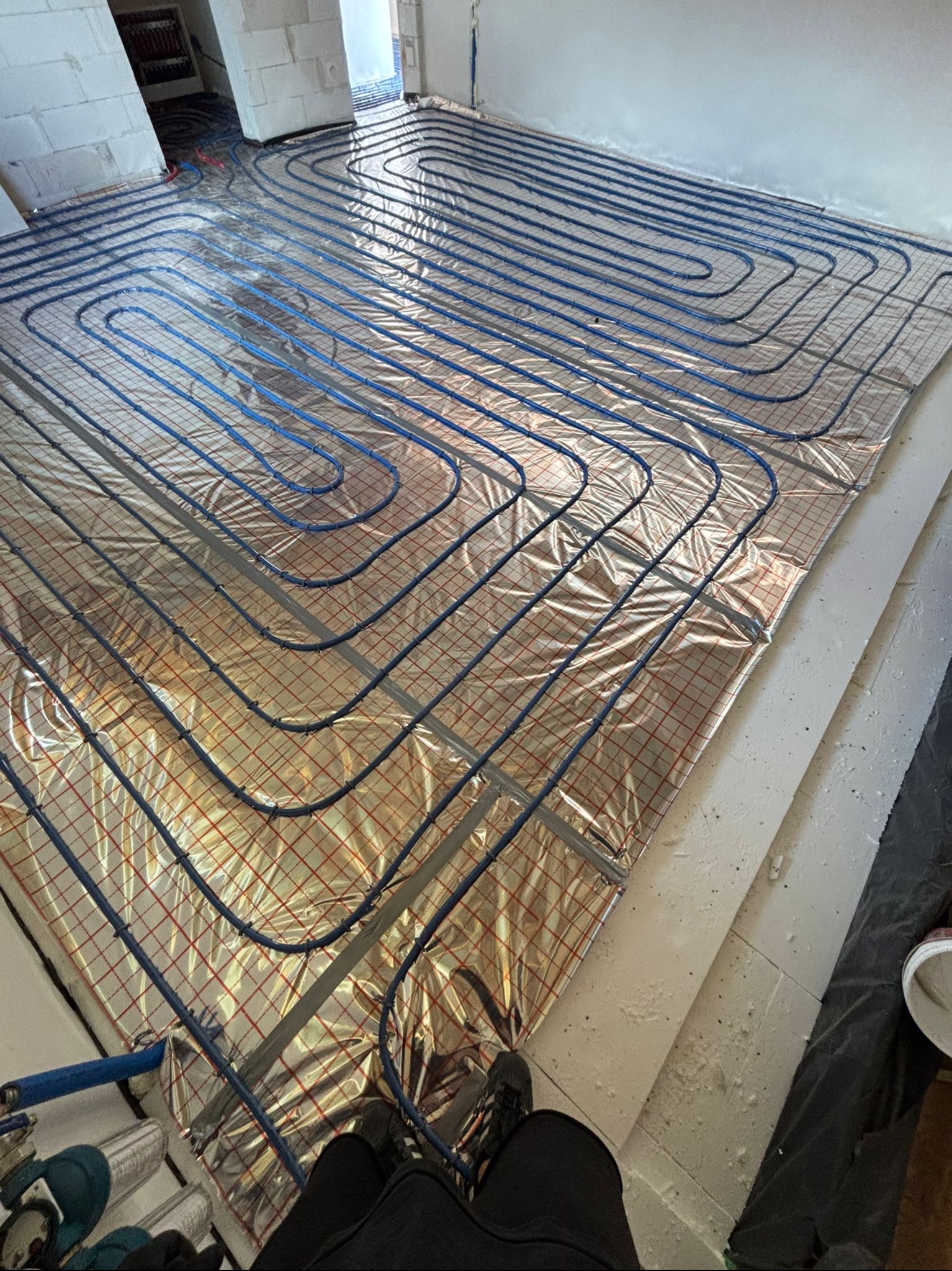 Rozłożona instalacja ogrzewania podłogowego na folii z siatką w surowym wnętrzu. Widoczne niebieskie rury ułożone w pętle i fragment osoby w kadrze.