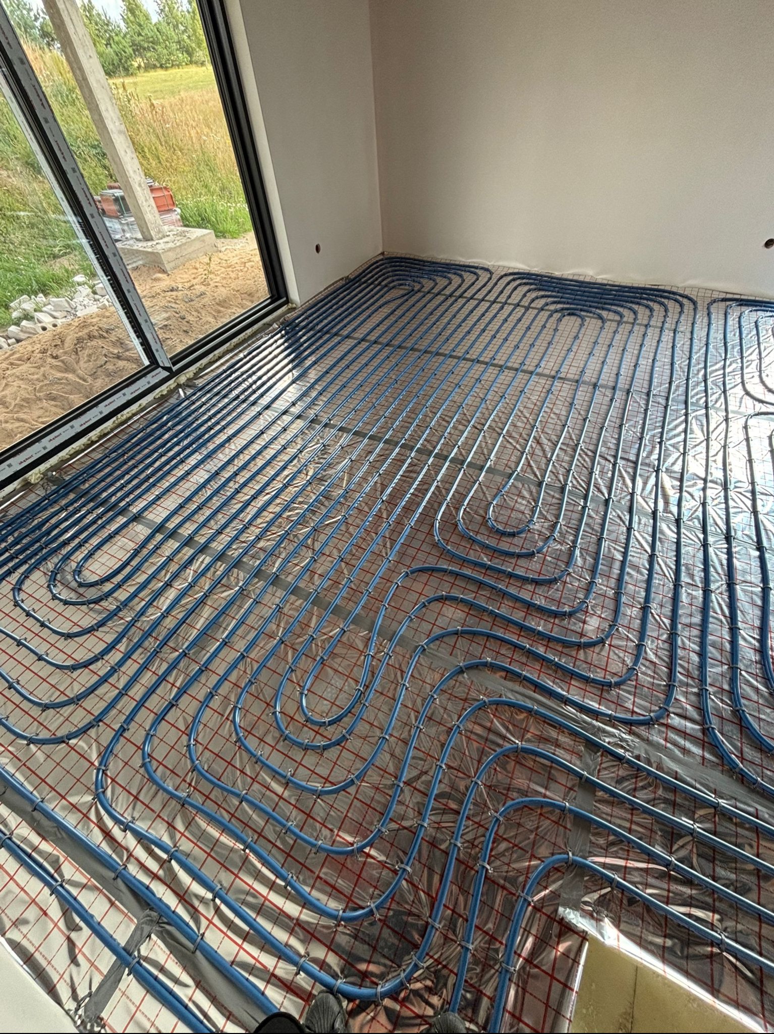 Niebieskie rury ogrzewania podłogowego rozłożone na srebrnej folii z czerwoną siatką w jasnym pomieszczeniu z dużym oknem na zewnątrz budynku w budowie.
