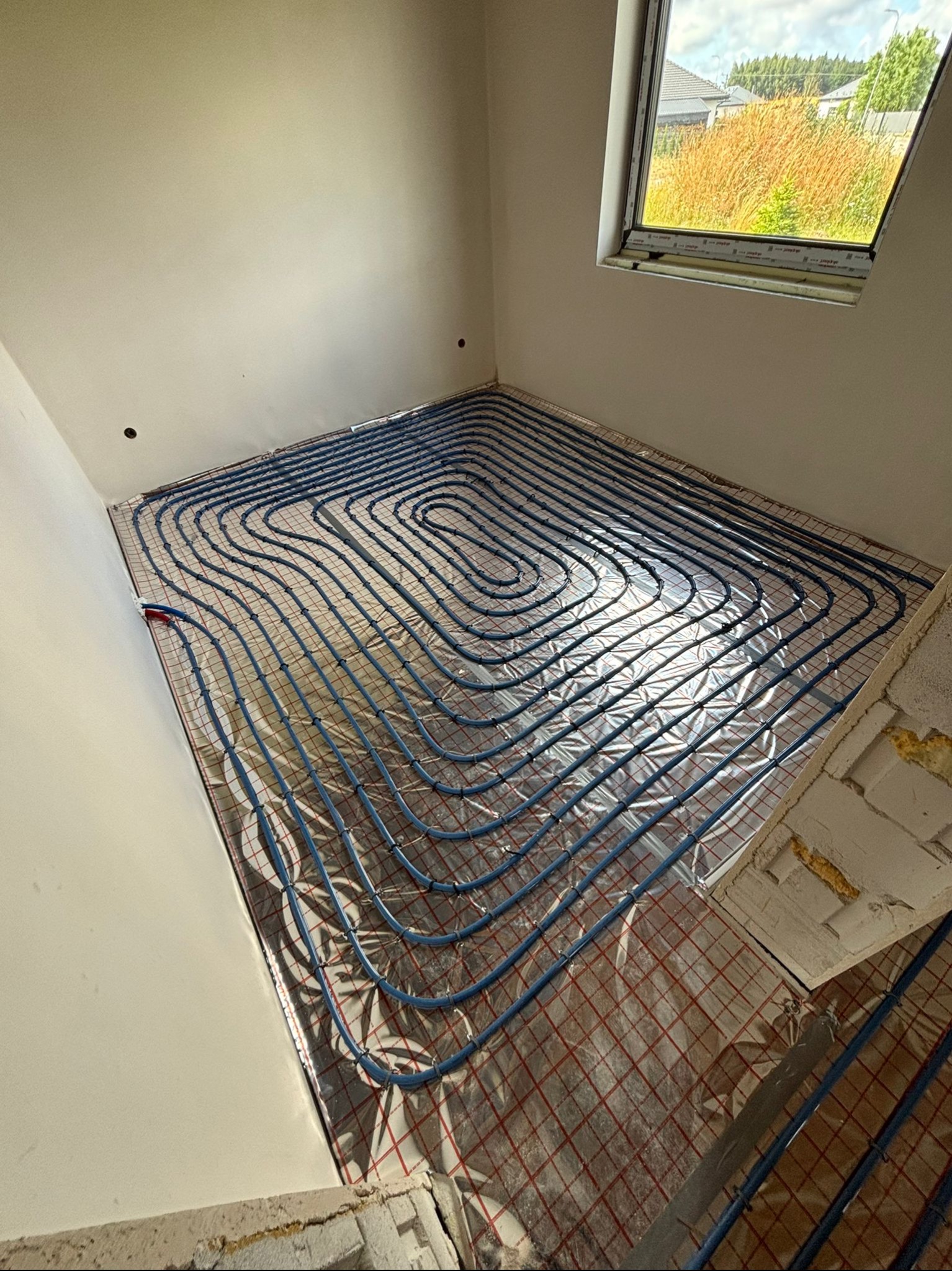 Niebieskie rury ogrzewania podłogowego ułożone spiralnie na srebrnej folii izolacyjnej w surowym wnętrzu z oknem, widok z góry na instalację przed wylewką.
