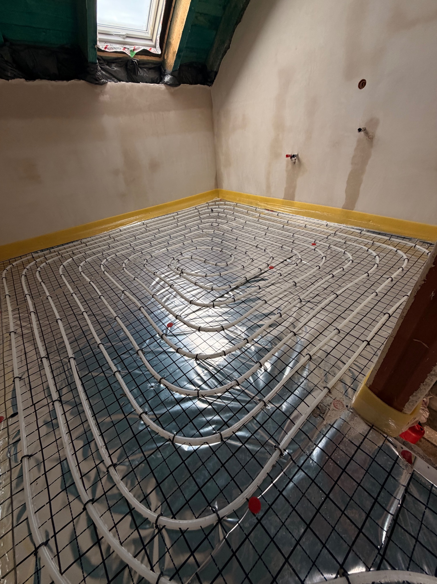 Białe rury ogrzewania podłogowego ułożone na folii izolacyjnej z czarną siatką, widok z góry, żółta taśma brzegowa, poddasze z oknem dachowym.