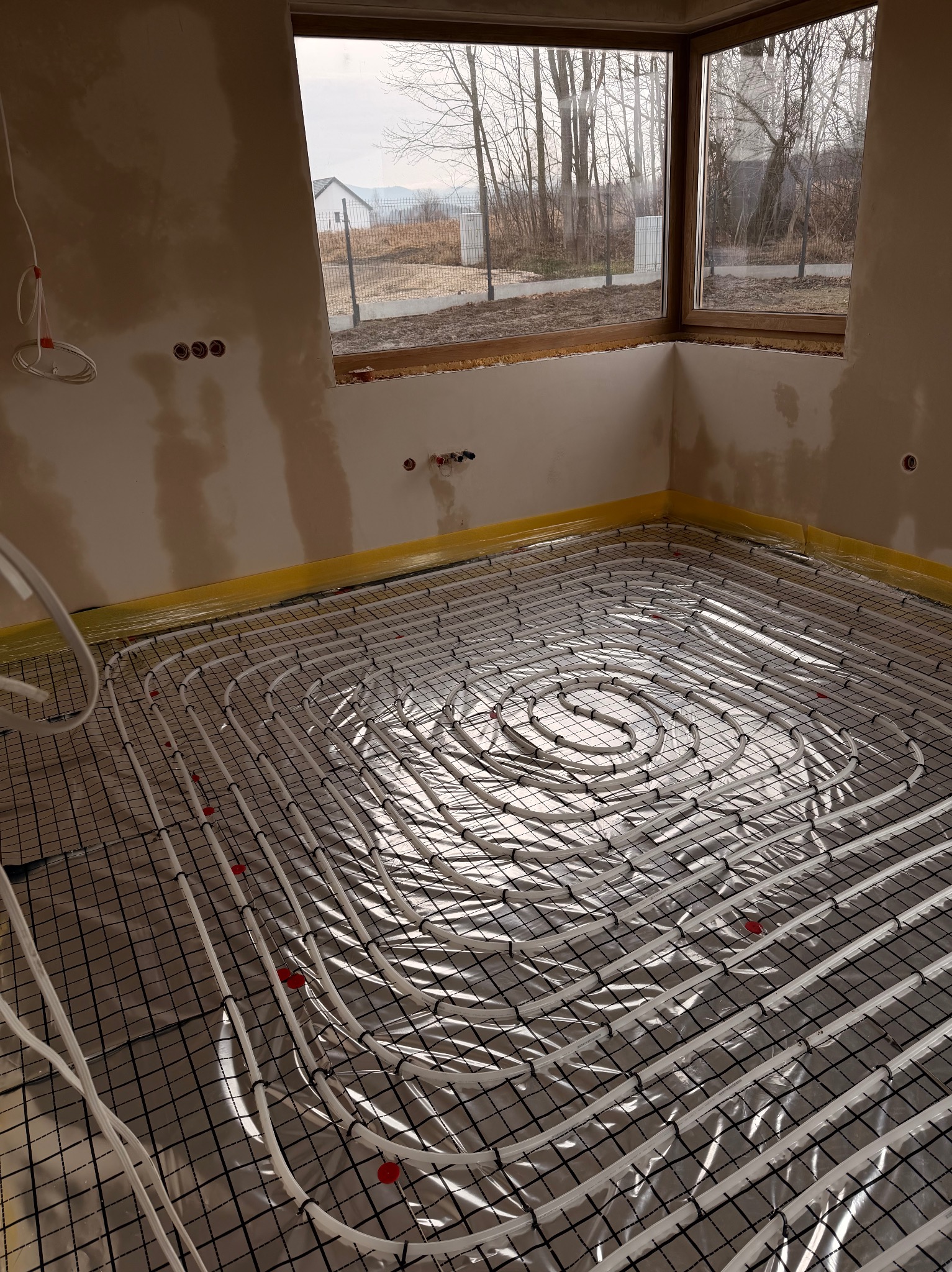 Ułożone rury ogrzewania podłogowego na folii, widoczne mocowania i okno z widokiem na zewnątrz. Ściany w stanie surowym, przygotowane do dalszych prac wykończeniowych.