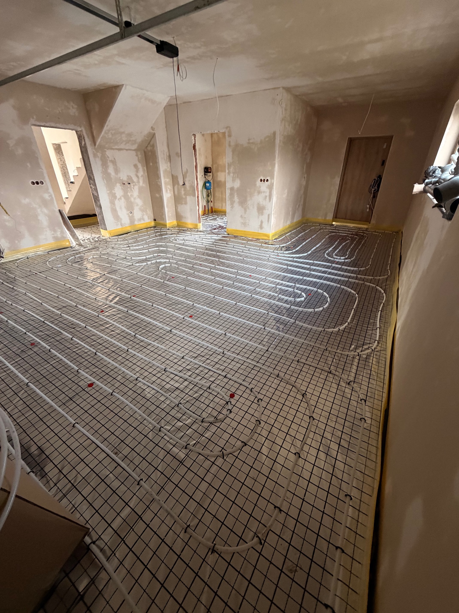 Białe rury ogrzewania podłogowego ułożone na czarnej siatce w surowym wnętrzu z żółtą taśmą przy ścianach. Widoczne drzwi i fragmenty instalacji.