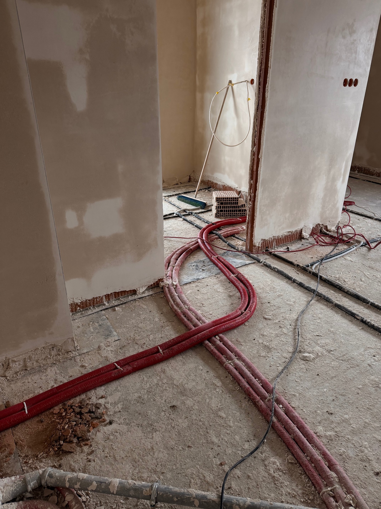Czerwone rury ogrzewania podłogowego ułożone na szarej posadzce w trakcie prac instalacyjnych wewnątrz budynku. Widoczne fragmenty ścian i otwory instalacyjne.