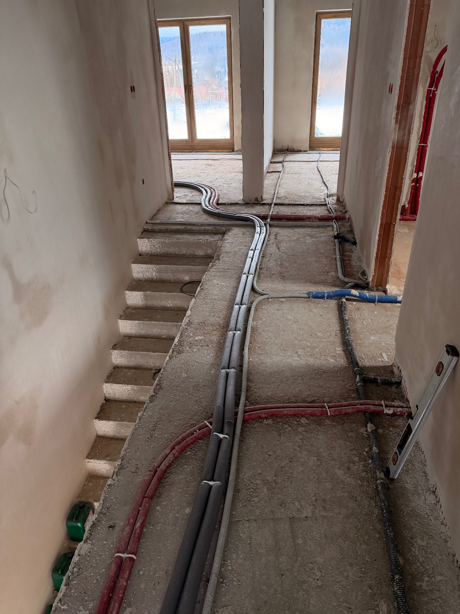 Instalacja ogrzewania podłogowego w surowym wnętrzu domu jednorodzinnego. Rury grzewcze ułożone na podłodze, widoczne schody i okna w tle. Poziomica przy ścianie.