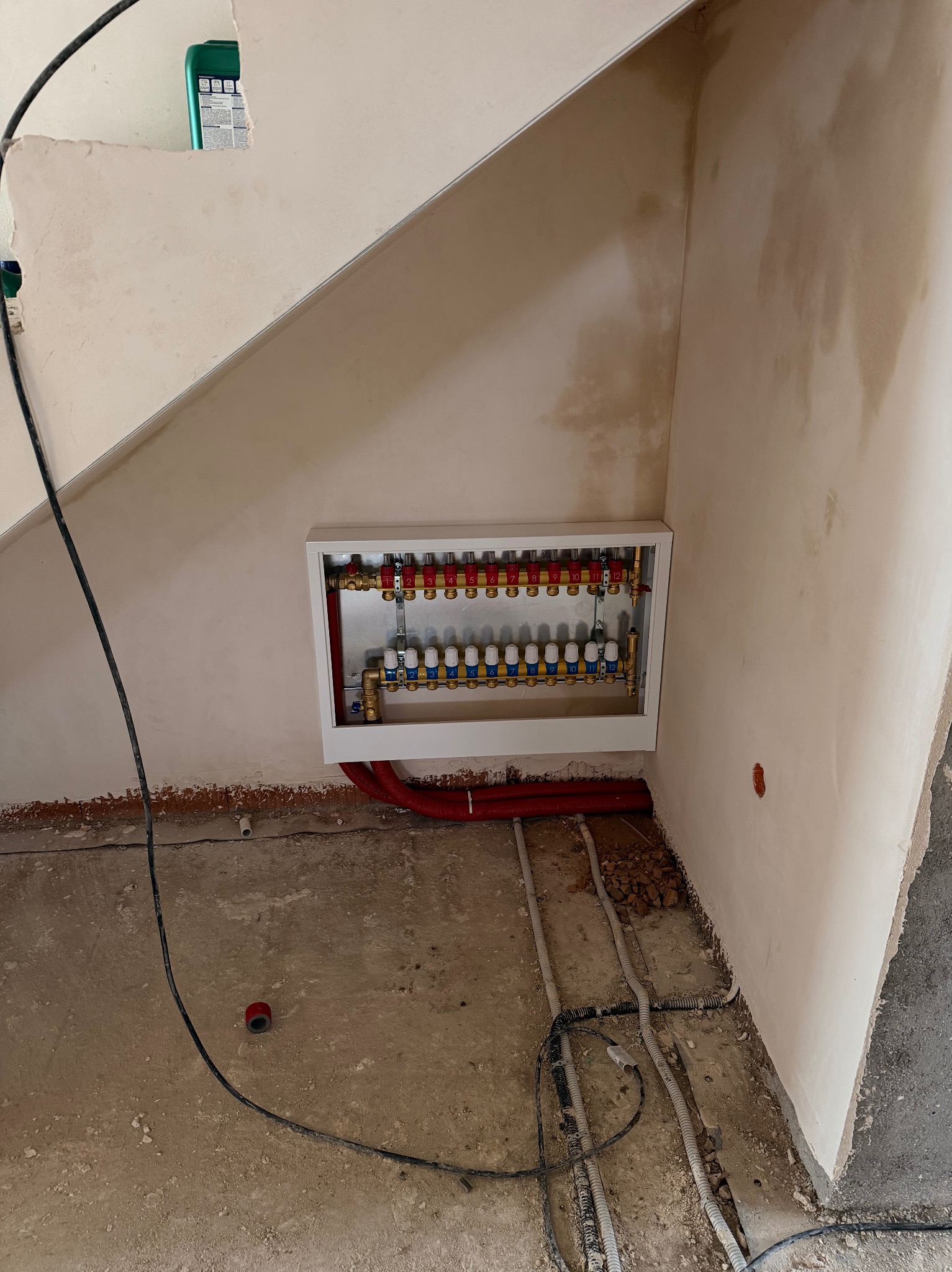 Rozdzielacz ogrzewania podłogowego w białej obudowie, z czerwonymi i niebieskimi zaworami, zamontowany na ścianie w surowym stanie. Widoczne rury i przewody na podłodze.