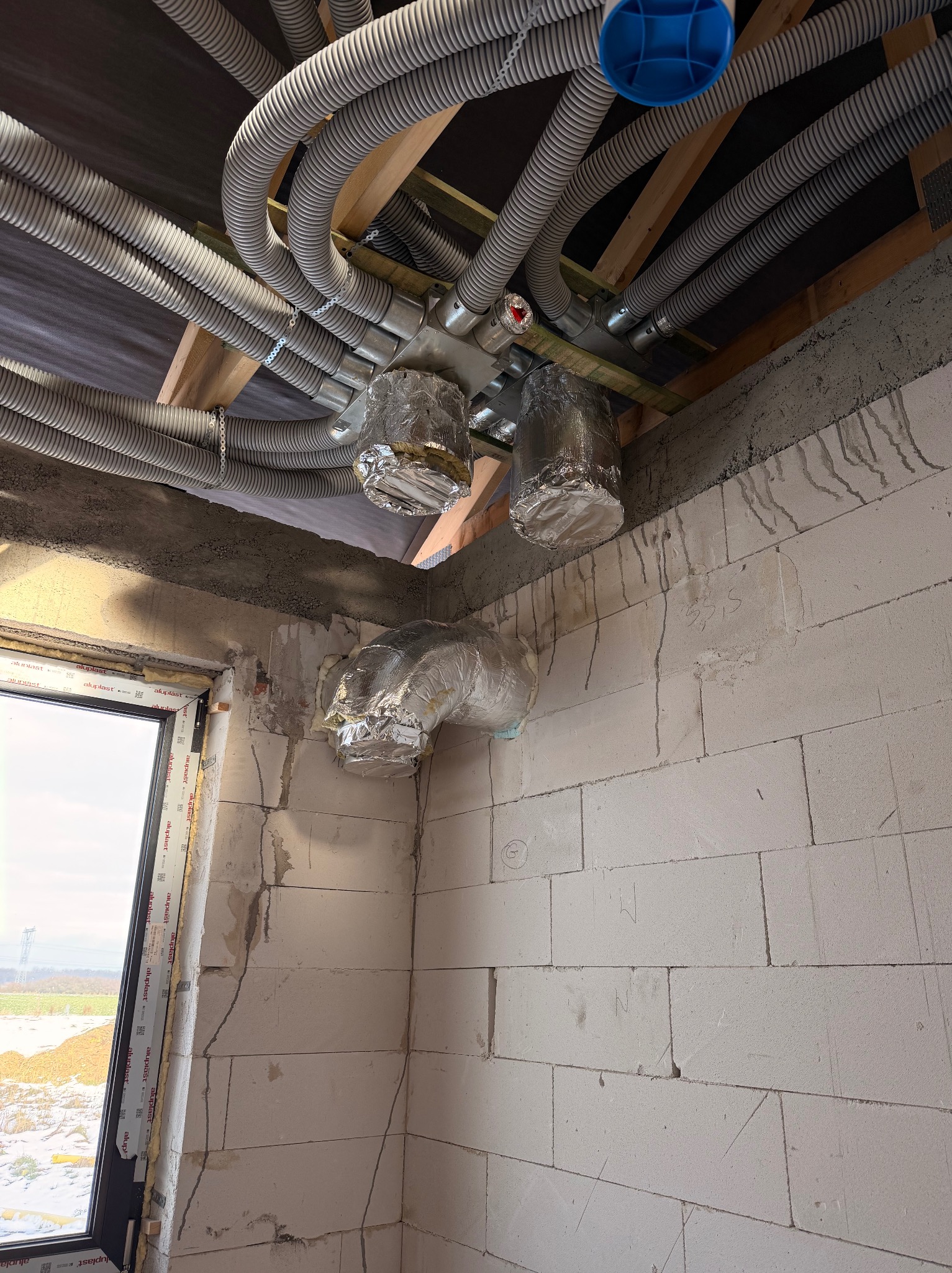 Szare, karbowane rury wentylacyjne pod sufitem, częściowo owinięte folią aluminiową, w surowym wnętrzu budynku z oknem. Widoczne elementy konstrukcyjne.