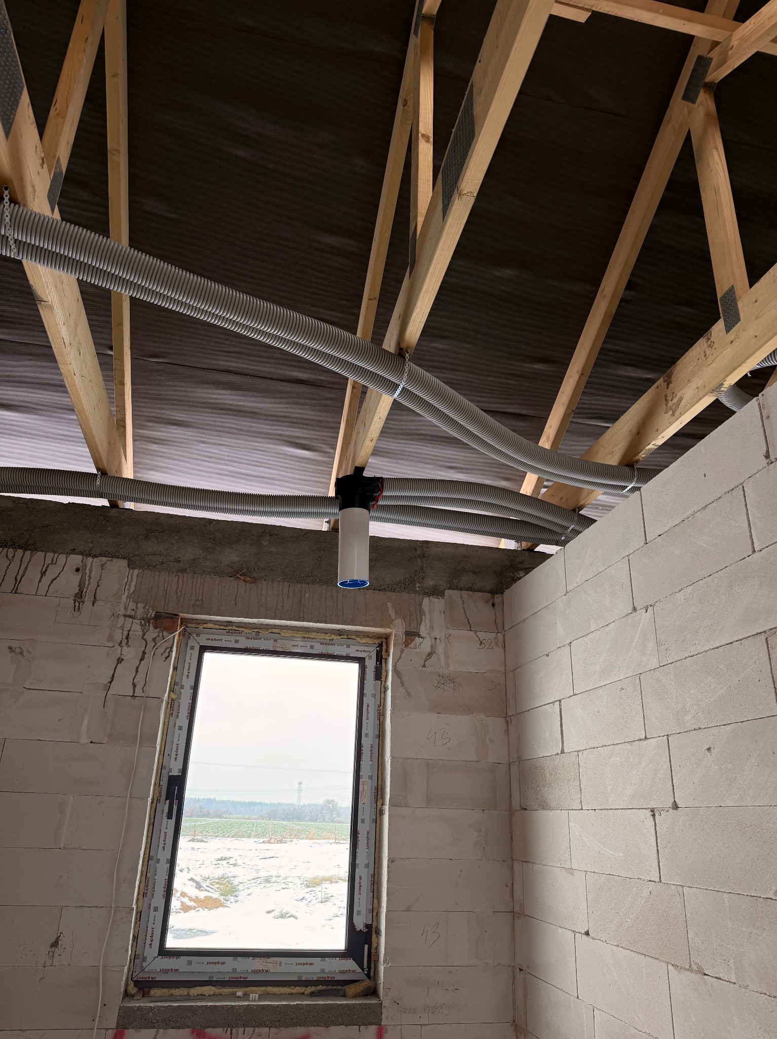 Szare, karbowane rury rekuperacji zamontowane pod drewnianą konstrukcją dachu w surowym wnętrzu budynku z oknem. Widoczny filtr rekuperatora.