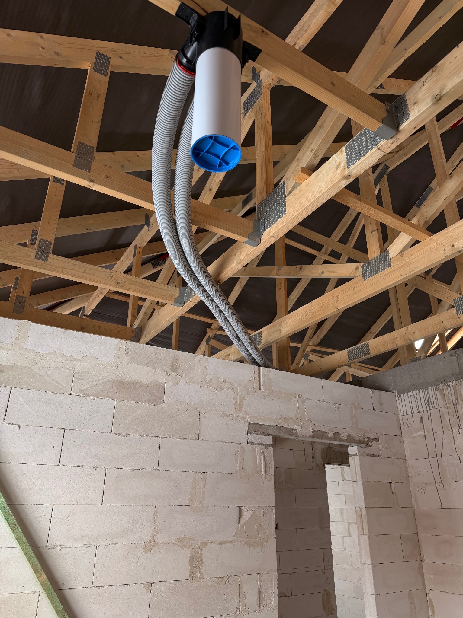 Montaż rekuperacji: biały moduł z niebieskim akcentem i szare rury pod drewnianą konstrukcją dachu w surowym wnętrzu z białych bloczków.