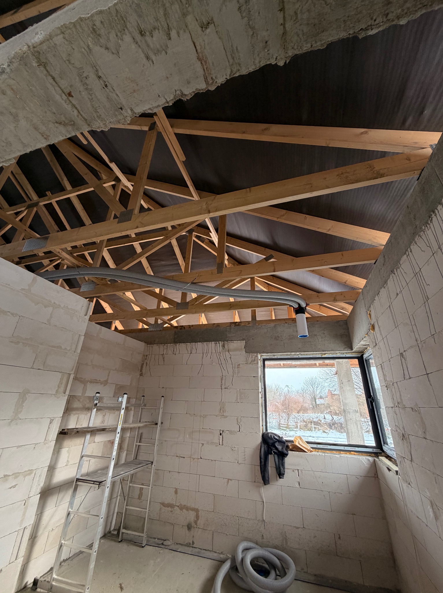 Montaż instalacji rekuperacji w surowym wnętrzu domu jednorodzinnego. Widoczne rury wentylacyjne pod drewnianą konstrukcją dachu i drabina.