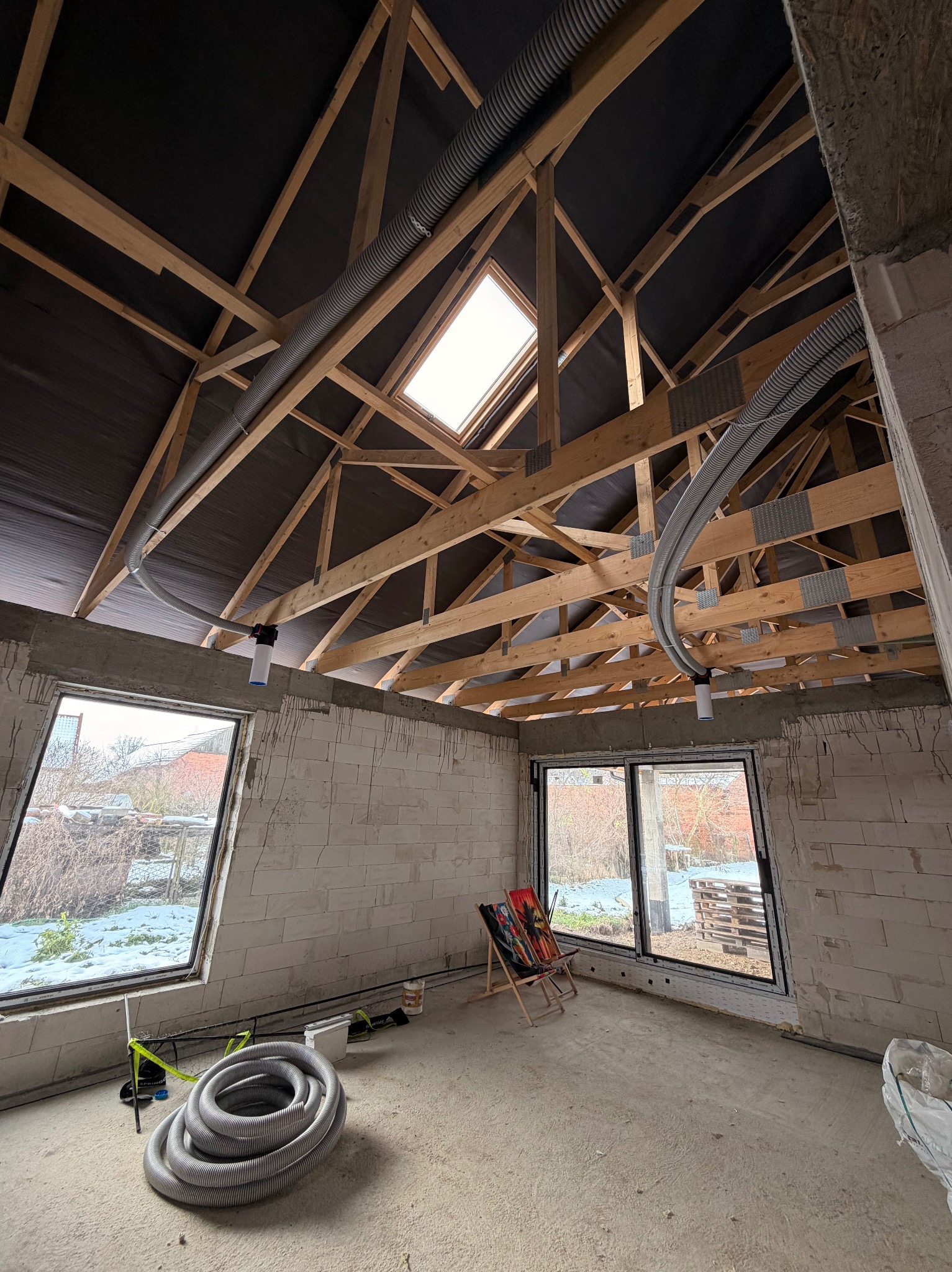 Surowy stan poddasza z widoczną konstrukcją dachu, oknem dachowym i częściowo zamontowaną instalacją wentylacyjną z rurami karbowanymi. W tle widok za oknem.