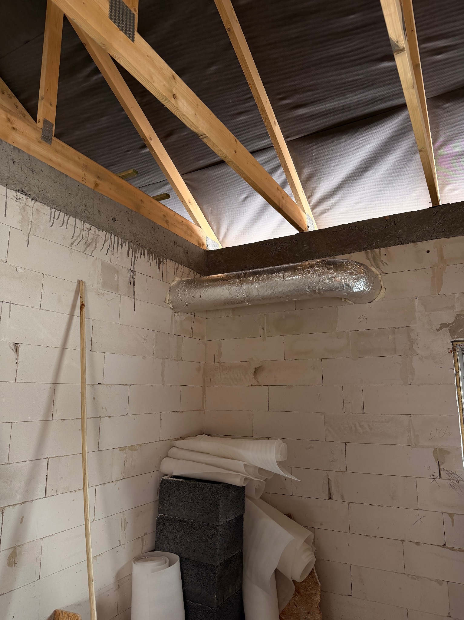 Wnętrze w trakcie budowy: surowa ściana z pustaków, widoczny fragment instalacji wentylacyjnej w srebrnej izolacji, drewniana konstrukcja dachu i materiały budowlane.