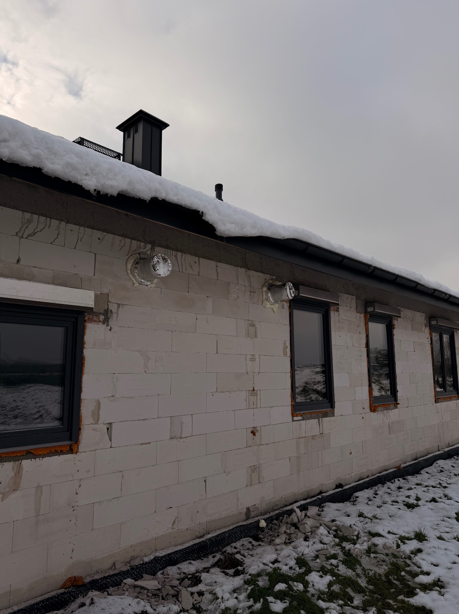 Budynek w trakcie montażu rekuperacji: widoczne otwory wentylacyjne na ścianie z białego bloczka, czarne okna i fragment dachu ze śniegiem. Zima.