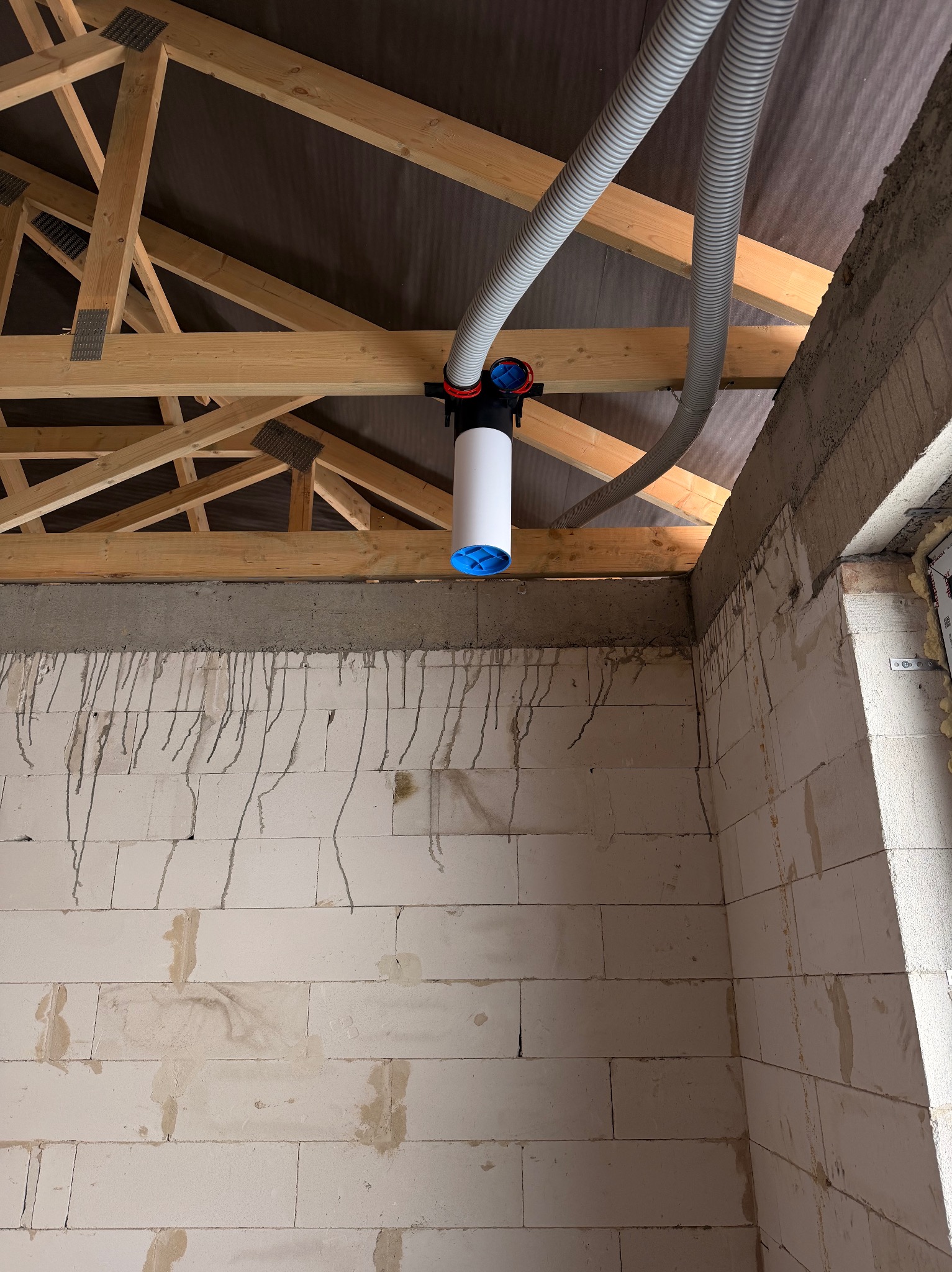 Fragment instalacji rekuperacji w surowym wnętrzu: biały filtr cylindryczny z niebieskimi akcentami, szare rury wentylacyjne, drewniana więźba dachowa i ściany z betonu komórkowego.