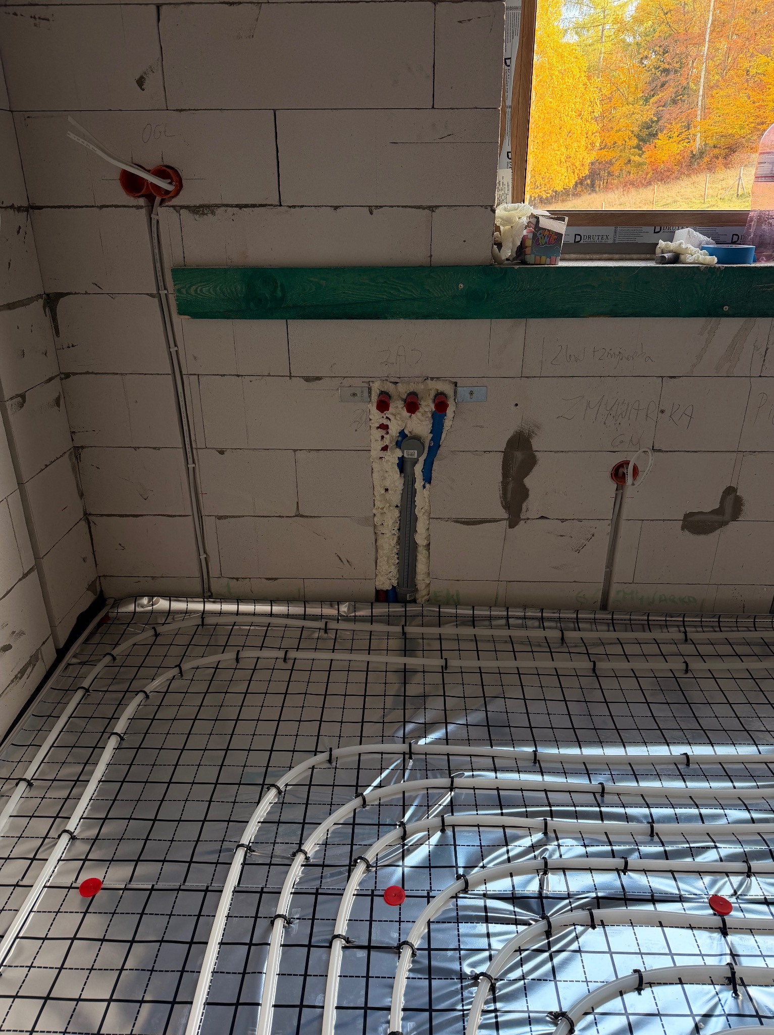 Białe rury ogrzewania podłogowego na folii refleksyjnej, widok z góry, instalacja w surowym stanie, okno z jesiennym widokiem w tle.