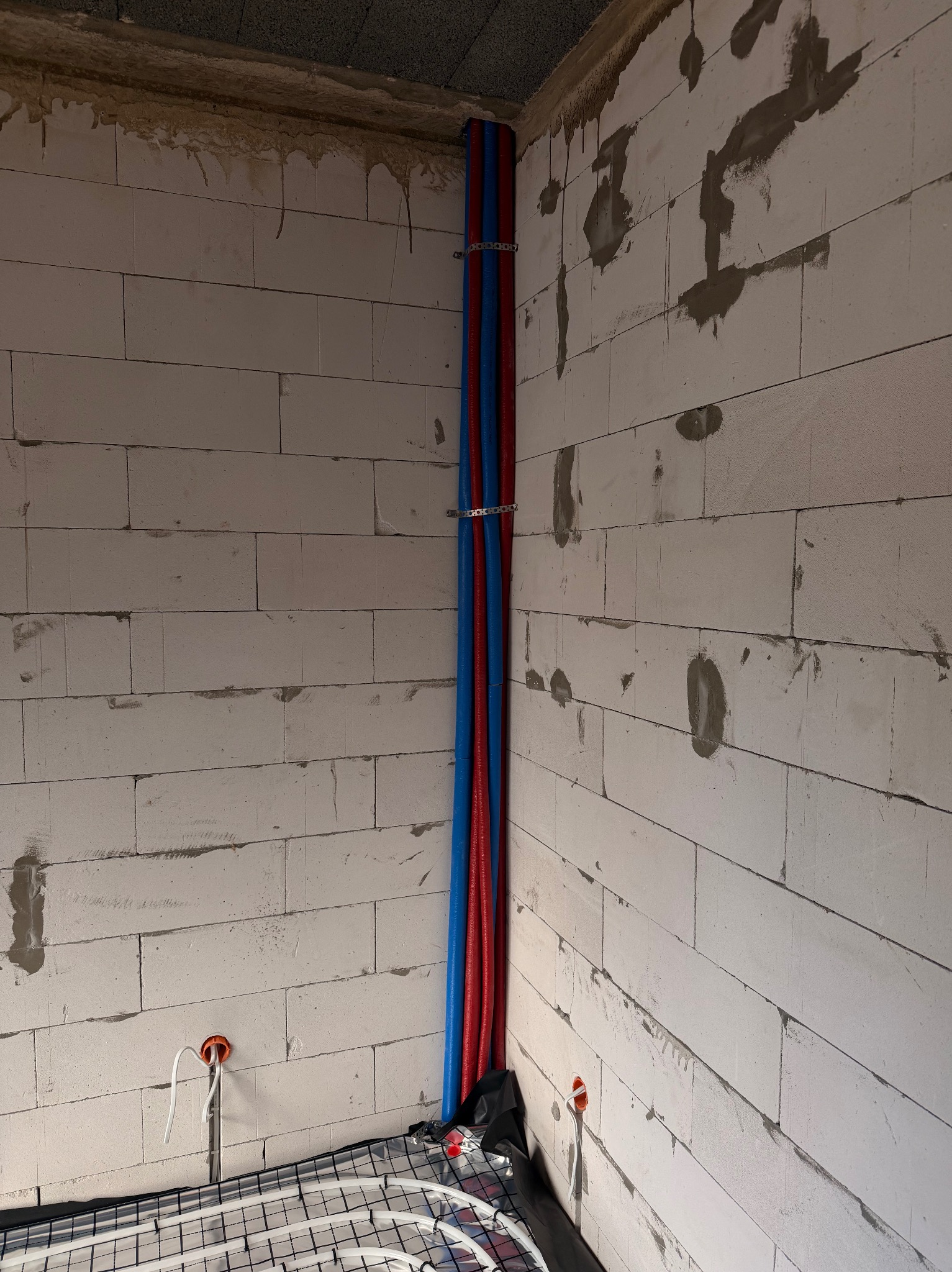 Instalacja ogrzewania podłogowego w surowym wnętrzu: rury grzewcze na folii, pionowe rury wodne (czerwona i niebieska) przy ścianie z betonu komórkowego.