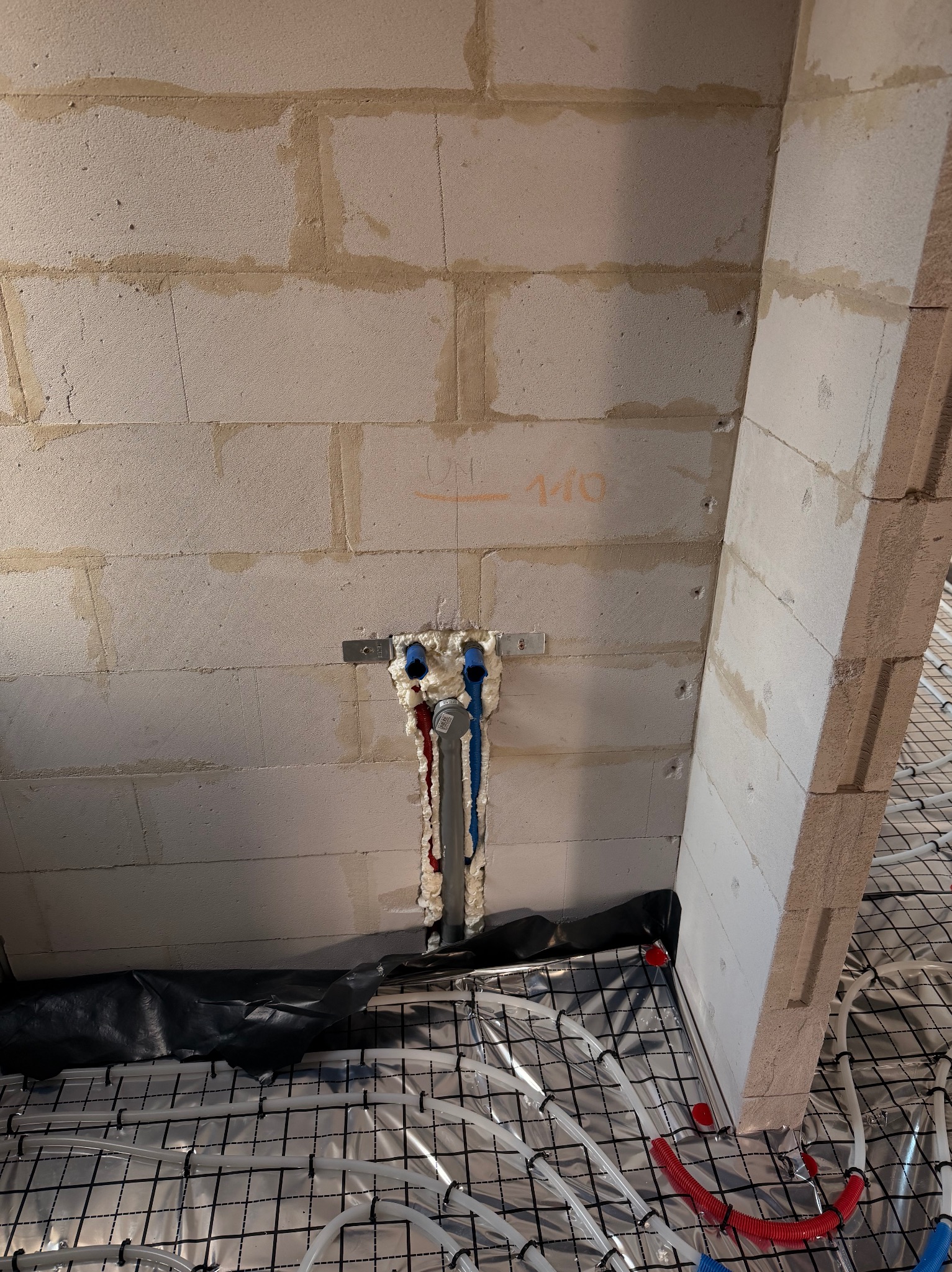 Instalacja ogrzewania podłogowego w trakcie montażu: rury grzewcze ułożone na izolacji, pionowe rury doprowadzające zabezpieczone pianką montażową przy ścianie z betonu komórkowego.