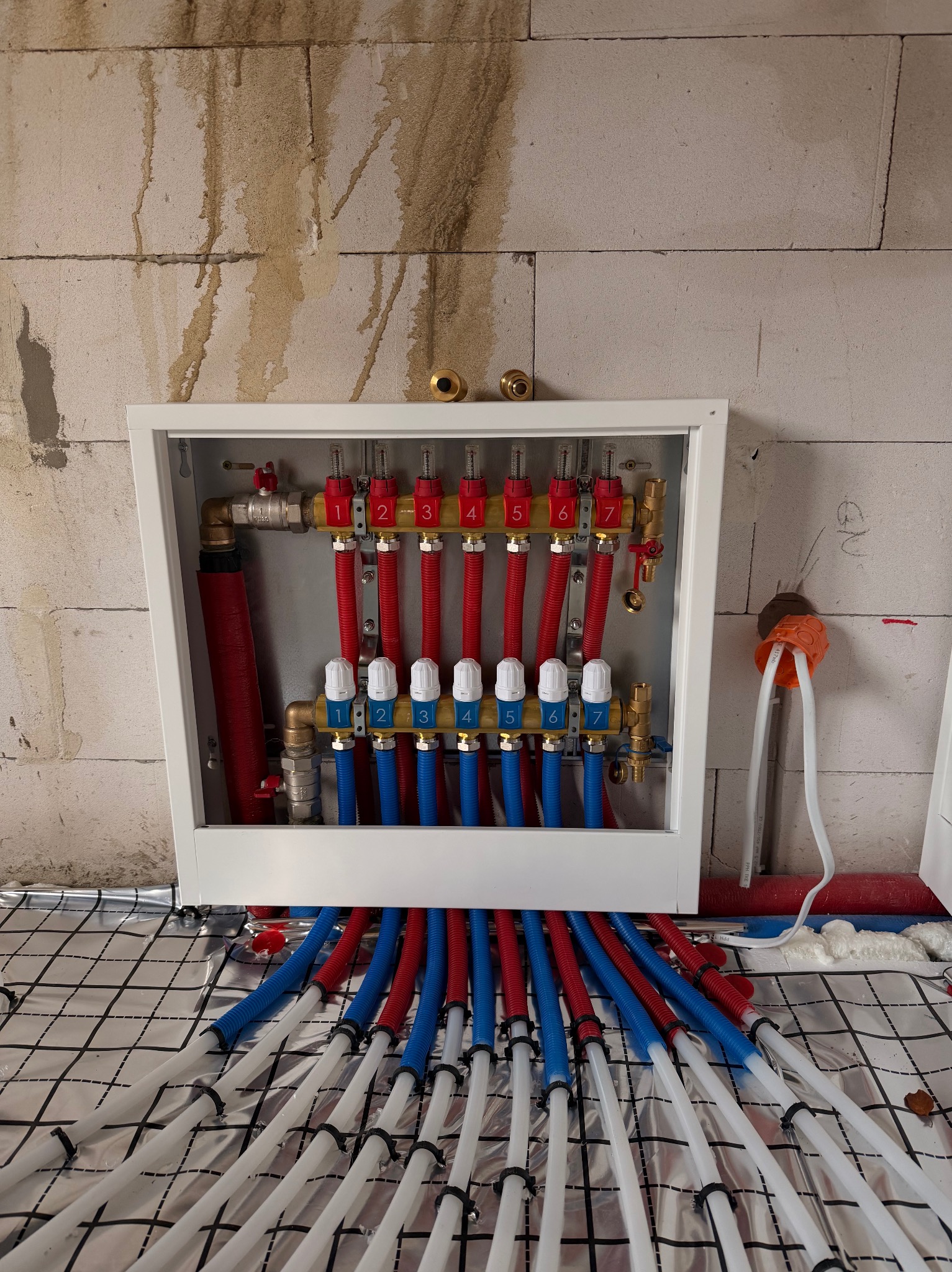 Rozdzielacz ogrzewania podłogowego z rurami w kolorach czerwonym i niebieskim, zamontowany na ścianie z bloczków, z widoczną folią izolacyjną na podłodze.