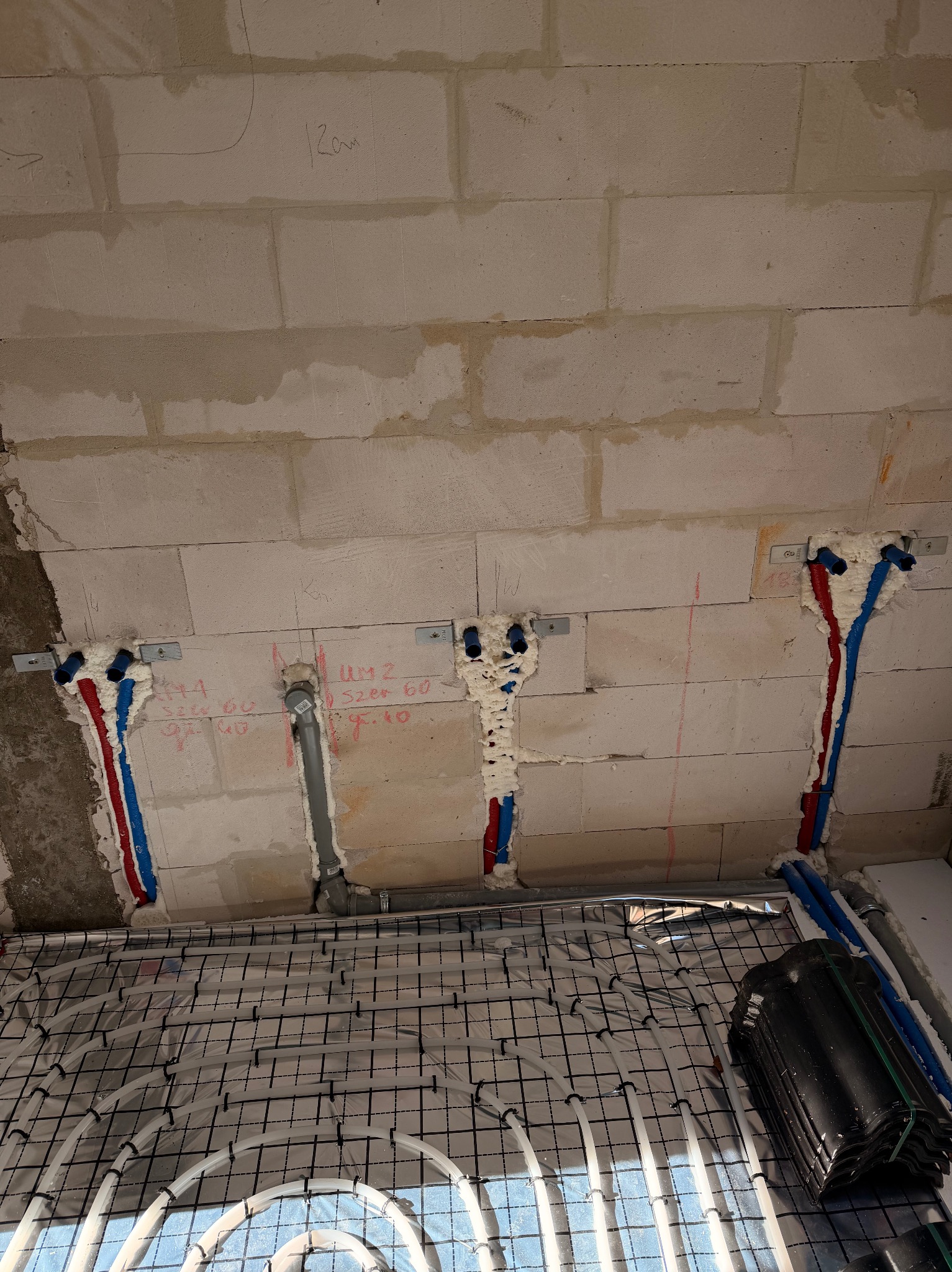 Montaż ogrzewania podłogowego: białe rury w siatce na podłodze, pionowe rury w ścianie z pianką montażową, czerwone i niebieskie. Ściana z bloczków.