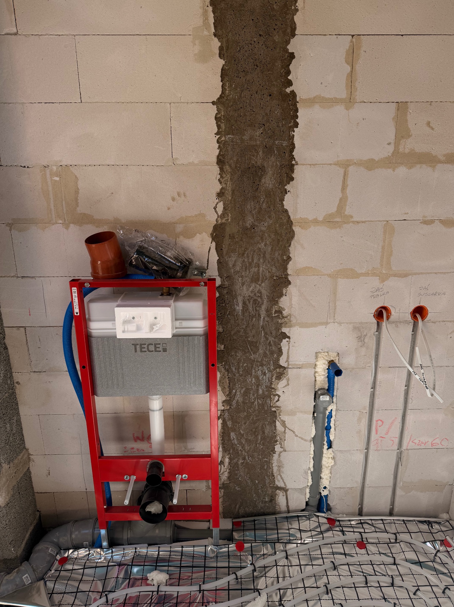 Czerwony stelaż podtynkowy TECE na tle ściany z betonu komórkowego, z widoczną instalacją ogrzewania podłogowego i rurami doprowadzonymi do ściany.