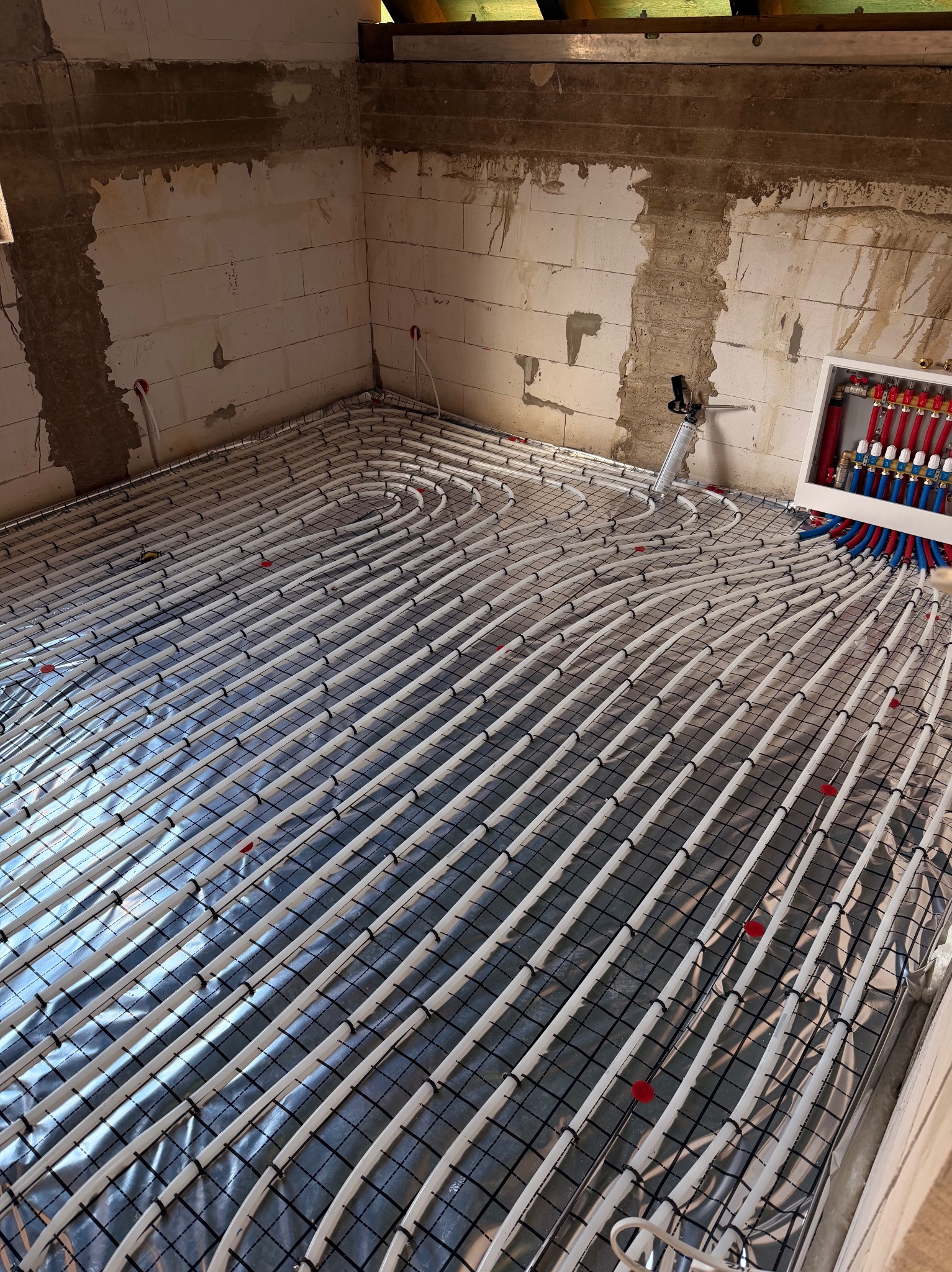 Białe rury ogrzewania podłogowego ułożone na folii, przymocowane do siatki, z rozdzielaczem w tle, w surowym wnętrzu budynku w trakcie budowy.