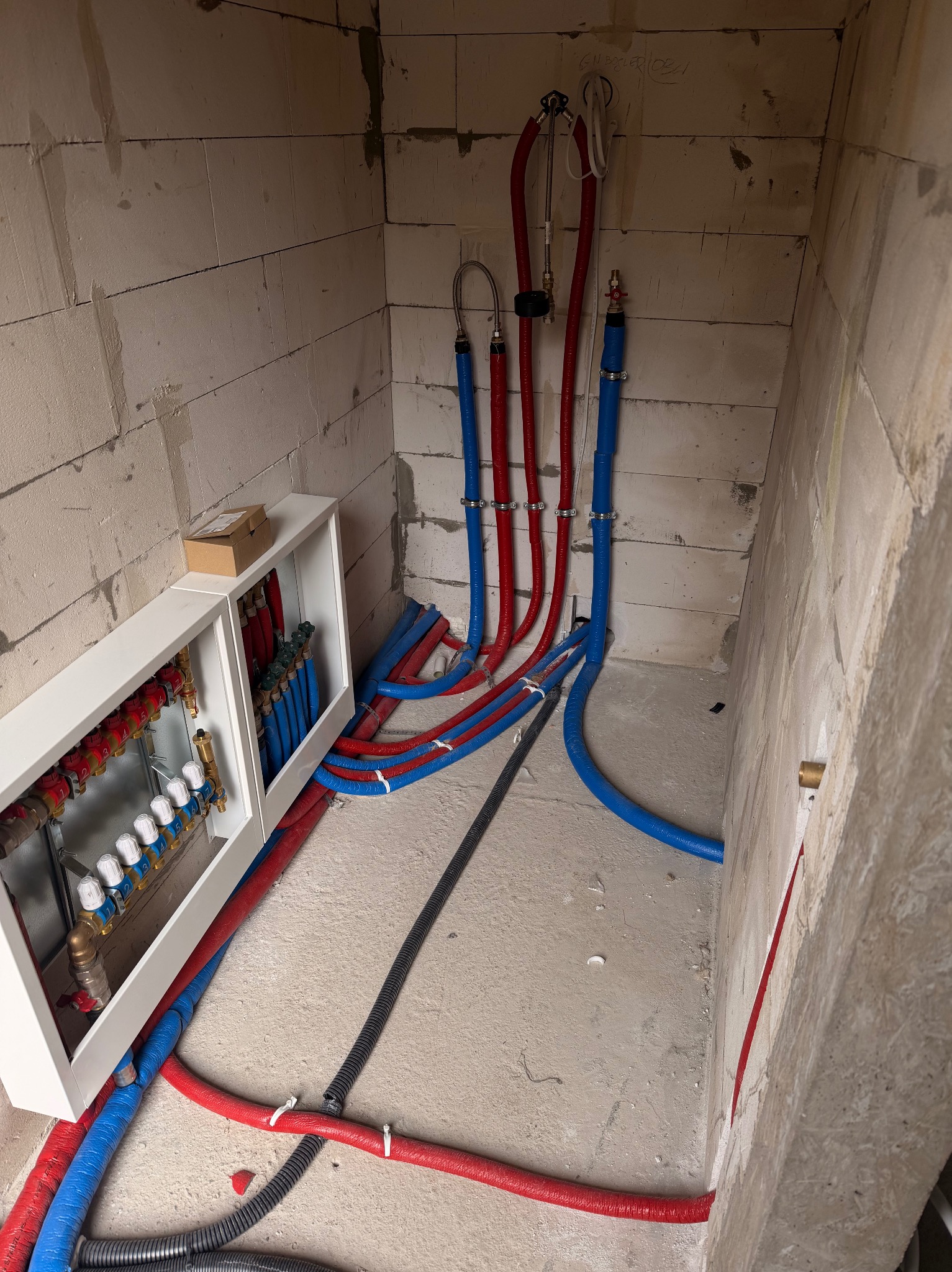 Rozdzielacz ogrzewania podłogowego z rurami w kolorach czerwonym i niebieskim, zamontowany w surowym pomieszczeniu z bloczków betonowych. Widoczne przewody i instalacja.