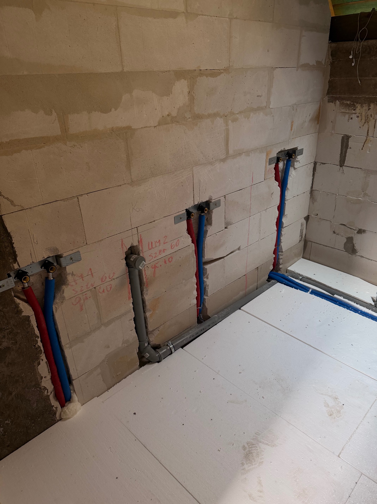 Instalacja wodna w ścianie z betonu komórkowego, rury PEX czerwone i niebieskie, szara rura kanalizacyjna, białe płyty styropianowe na podłodze, etap prac instalacyjnych.