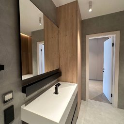 Kult Interiors - Nowoczesna łazienka z szafką pod umywalkę w odcieniach szarości i bieli, drewniana szafa wnękowa i duże lustro z czarną półką. Minimalistyczny design, stonowana kolorystyka.