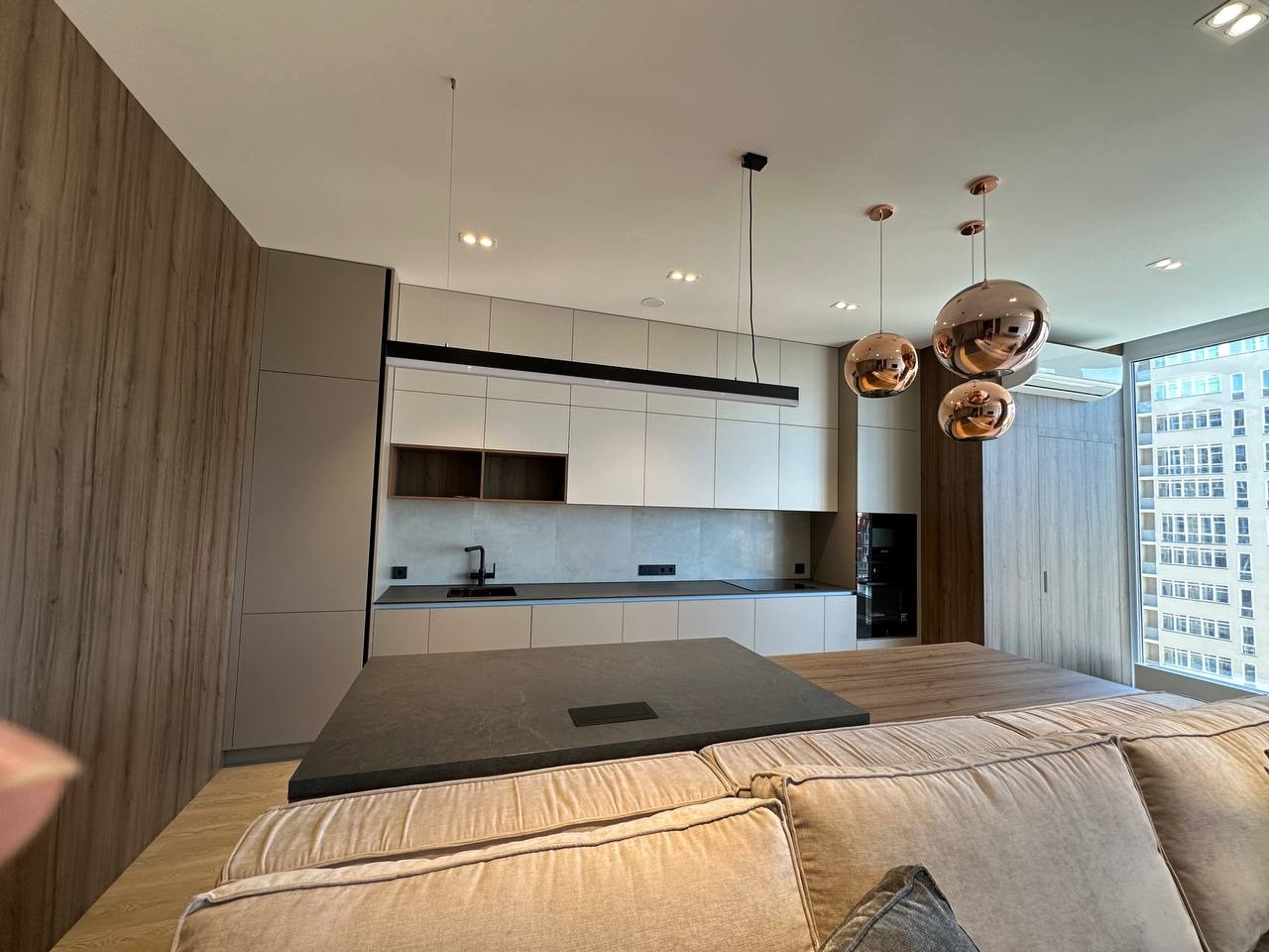 Nowoczesna kuchnia z wyspą, beżowe szafki i drewniane akcenty, oświetlenie kuliste, widok zza sofy. Minimalizm i funkcjonalność w jednym.