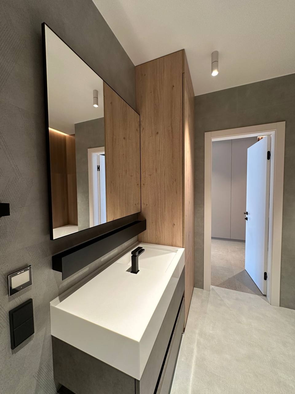 Nowoczesna łazienka z szafką pod umywalkę w odcieniach szarości i bieli, drewniana szafa wnękowa i duże lustro z czarną półką. Minimalistyczny design, stonowana kolorystyka.