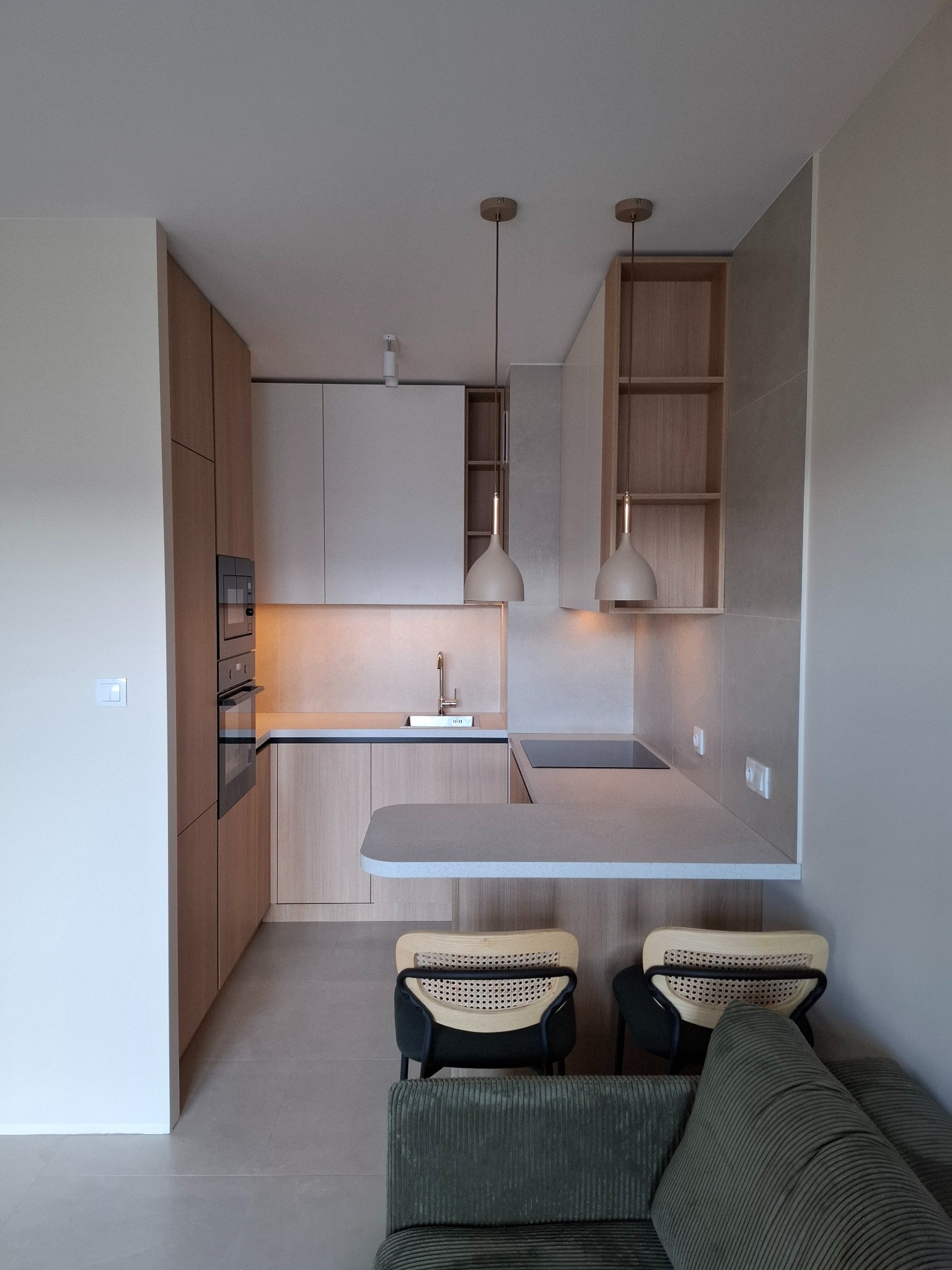 Nowoczesna kuchnia z zabudową na wymiar w odcieniach beżu i szarości, oświetlona wiszącymi lampami. Widoczne krzesła barowe i fragment sofy.