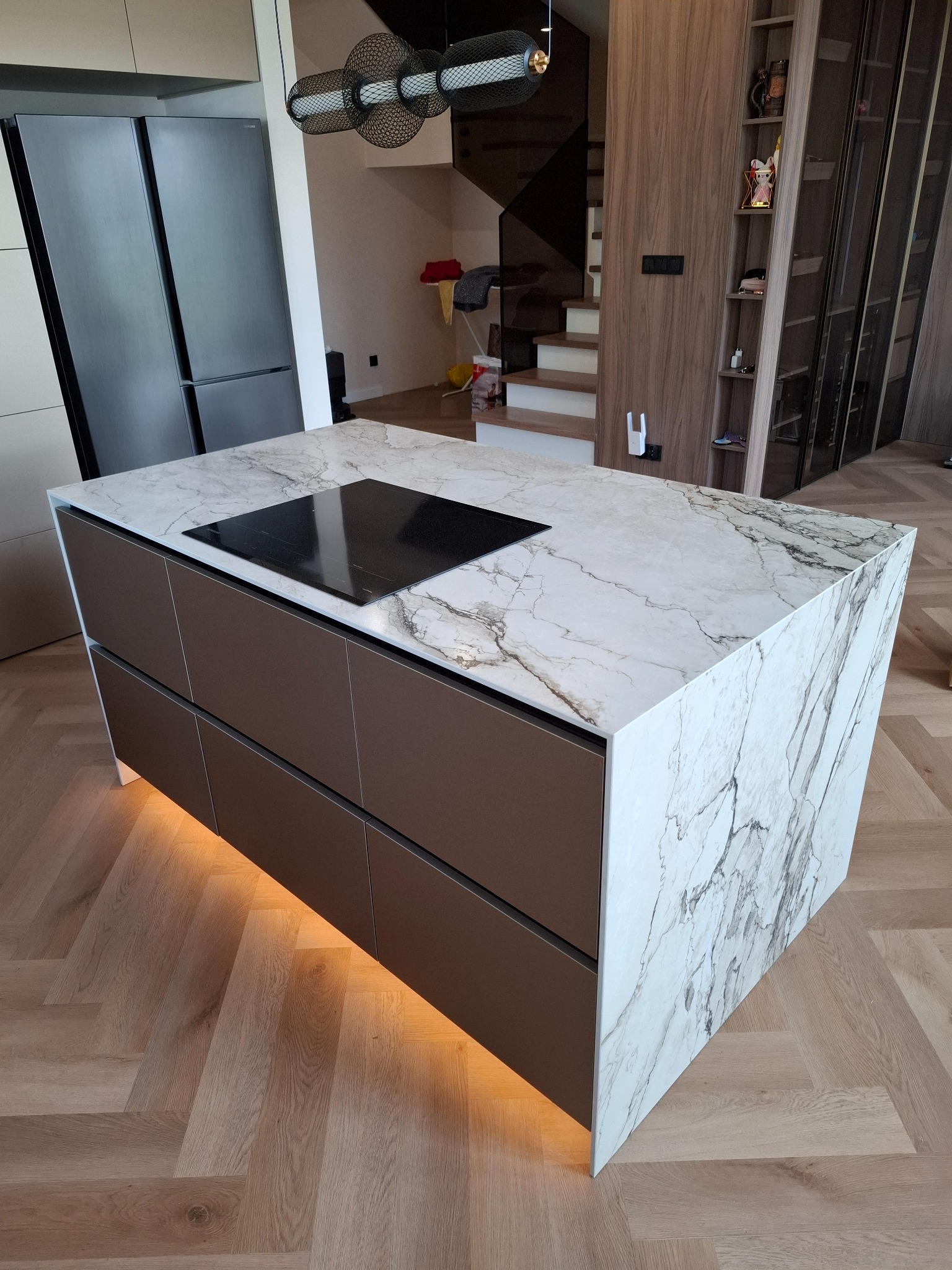 Nowoczesna wyspa kuchenna z marmurowym blatem i płytą indukcyjną, brązowe fronty szafek z subtelnym podświetleniem LED, parkiet w jodełkę.