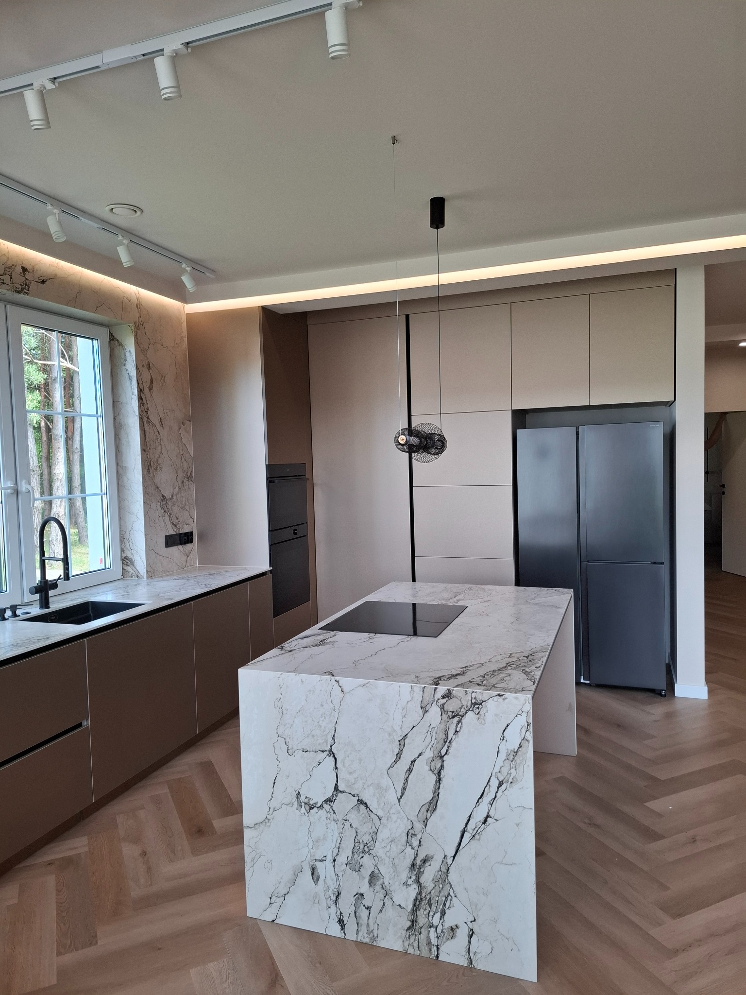 Nowoczesna kuchnia z marmurową wyspą i beżowymi szafkami, czarny zlew przy oknie, parkiet w jodełkę, oświetlenie punktowe i wisząca lampa, lodówka w zabudowie.